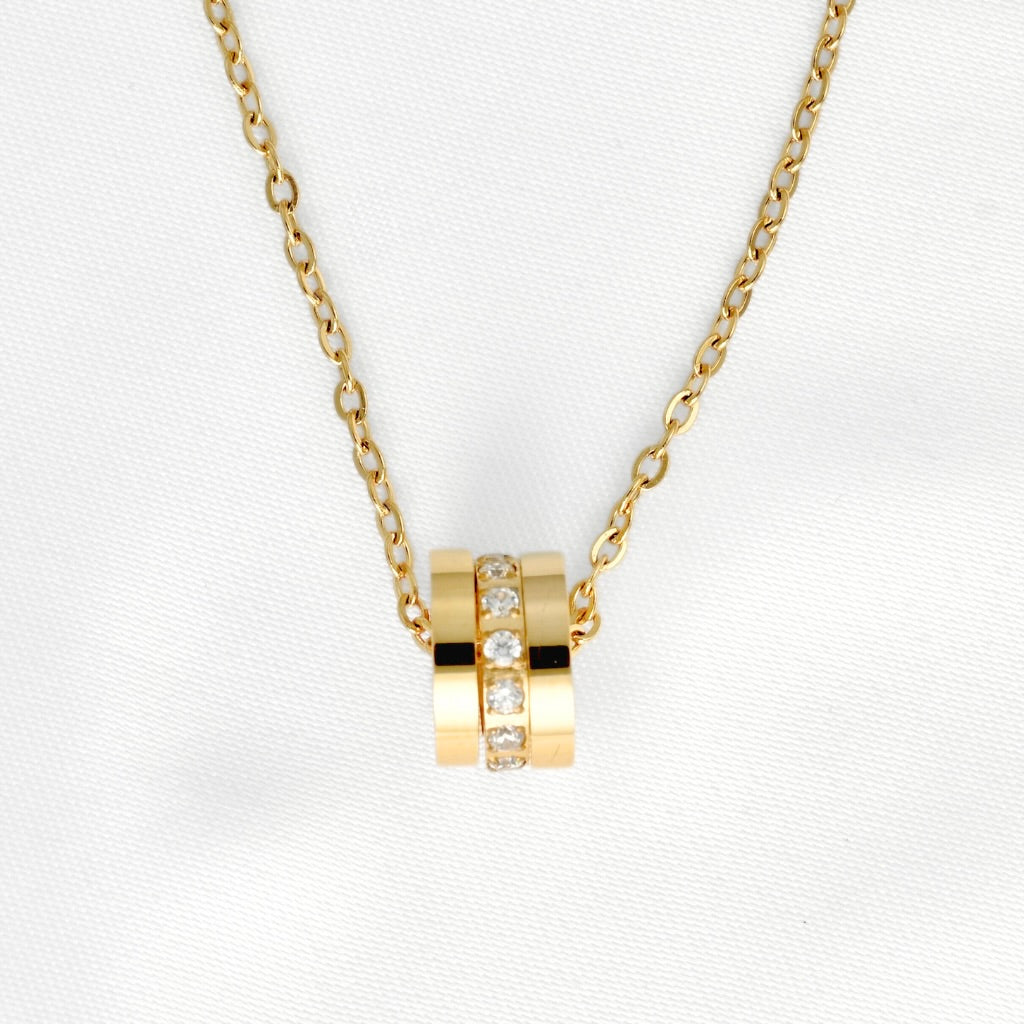 A gold chain necklace with a pendant consisting of three stacked gold rings, the middle one one adorned with small clear stones, on a textured white background.