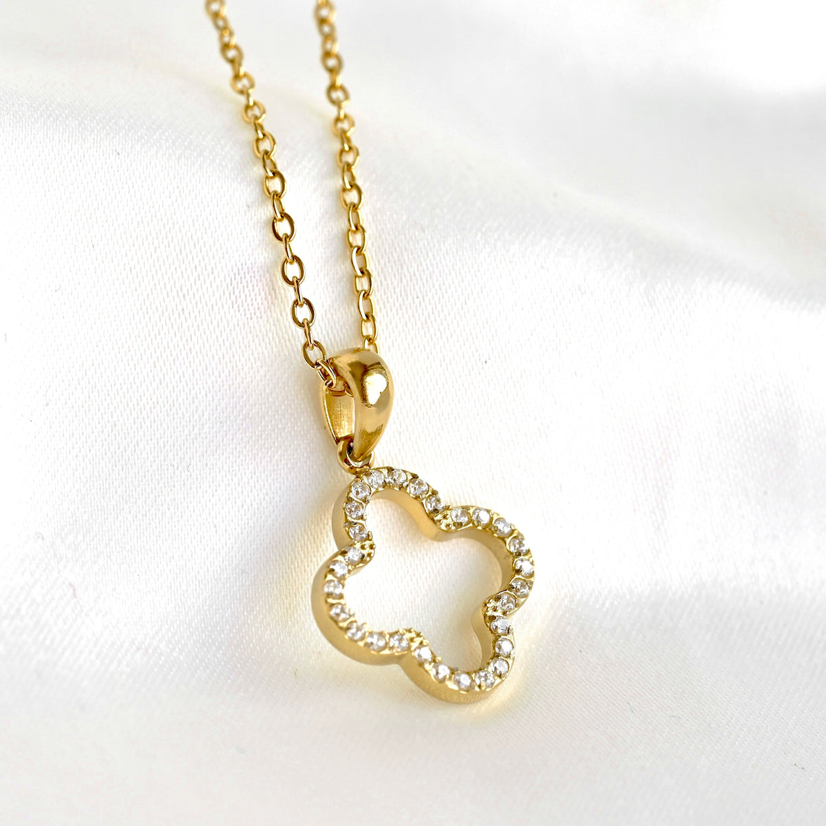 A gold necklace with a clover-shaped pendant adorned with small clear crystals, displayed on a soft white fabric background.