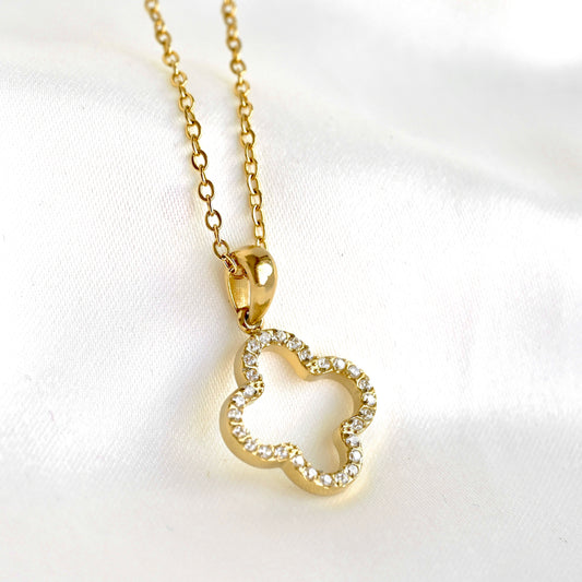 A gold necklace with a clover-shaped pendant adorned with small clear crystals, displayed on a soft white fabric background.