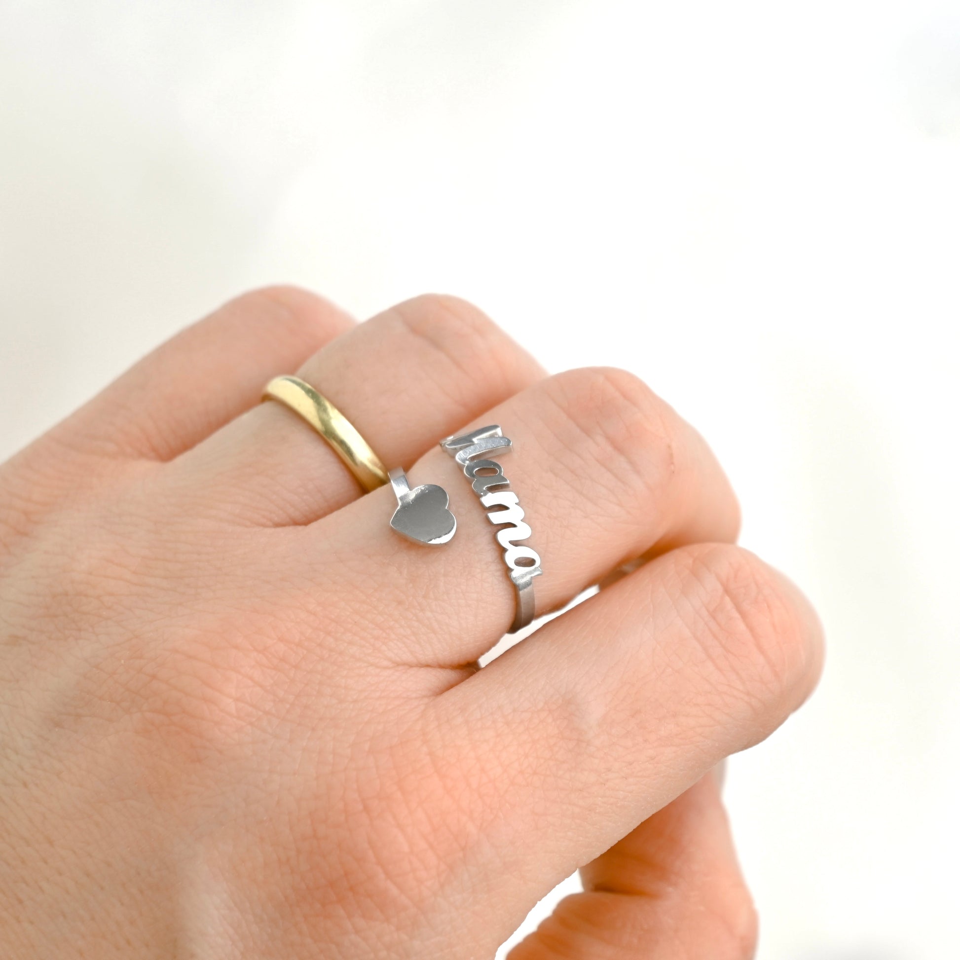 A close-up of a hand wearing two rings: a simple gold band and a silver mama heart 