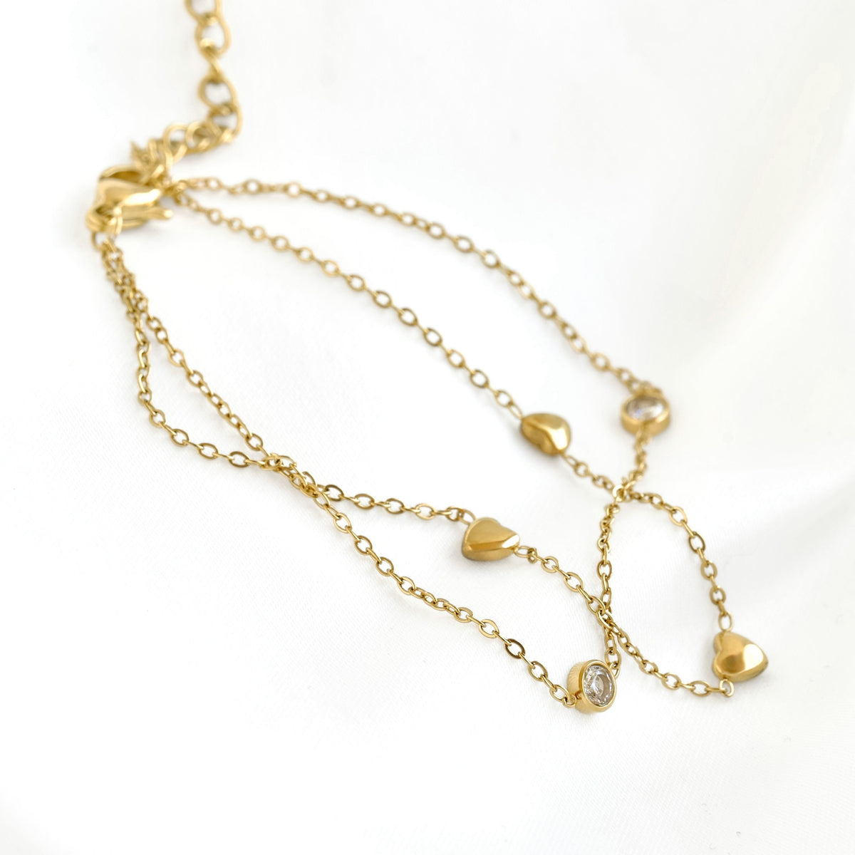 A delicate gold chain bracelet featuring multiple draped chains with small gold heart charms and a clear round gemstone accent, displayed on a white fabric background.