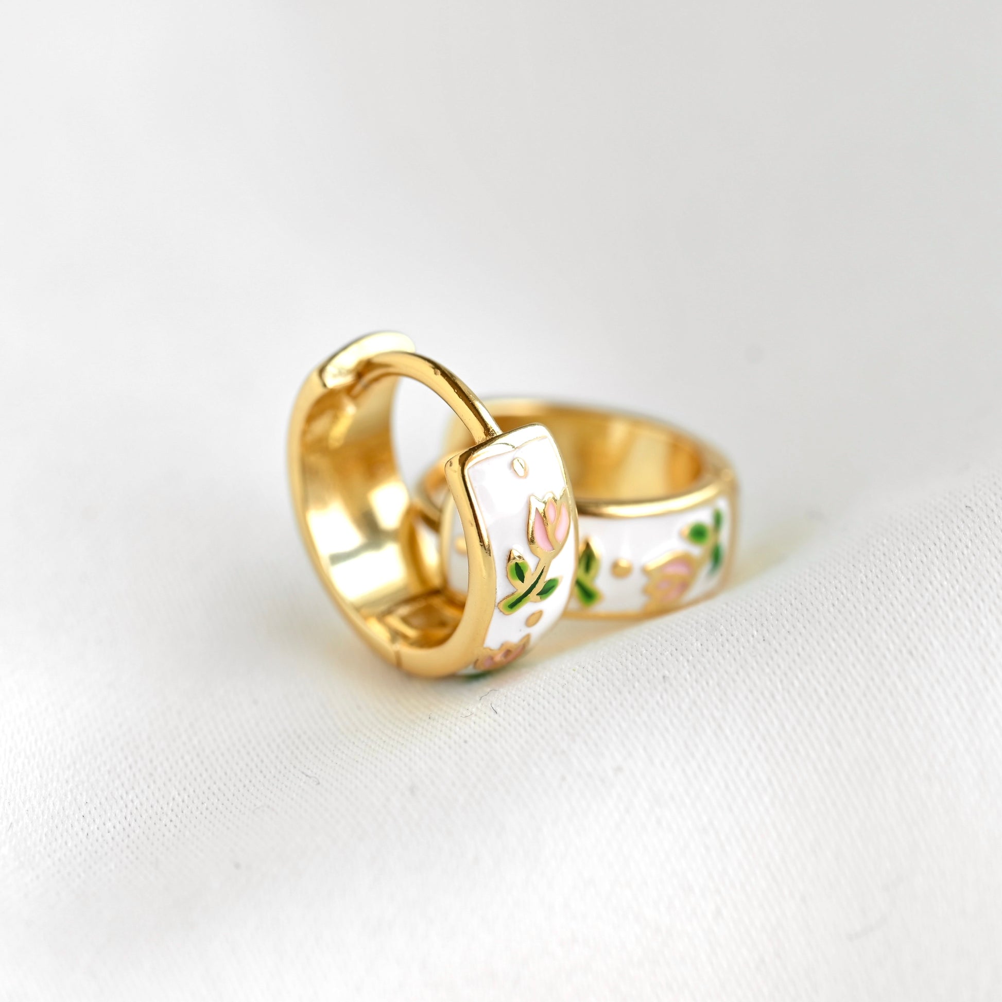 A gold-plated huggie ring with a white enamel inlay featuring a delicate pink and green floral pattern, resting on a white textured fabric background.