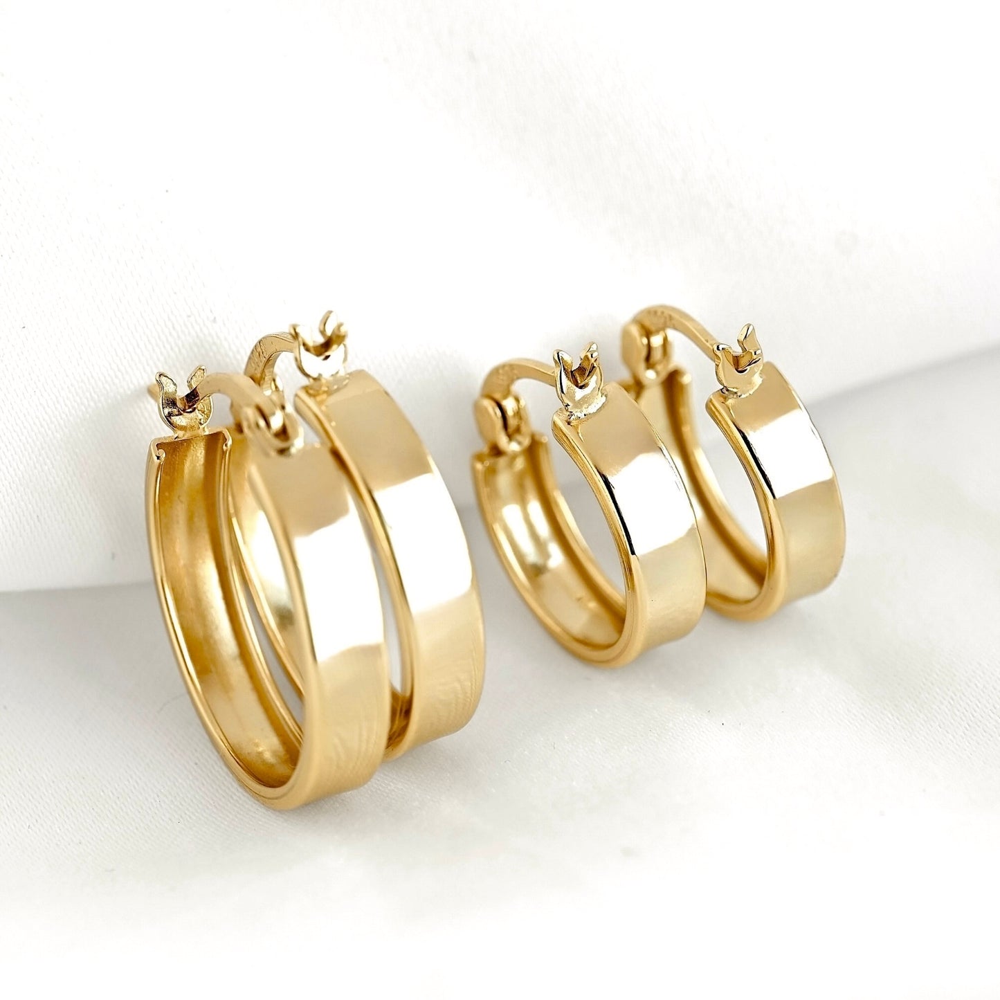 A pair of polished gold hoop earrings with small latch back closure, displayed on a soft white fabric background.