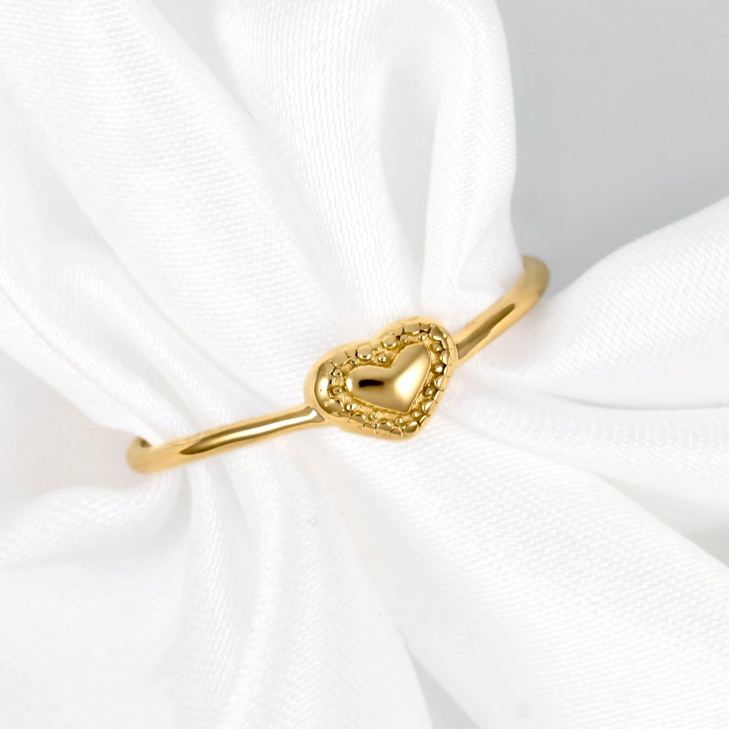 A gold ring with a milgrain heart-shaped  charm, displayed on a white fabric ring holder.