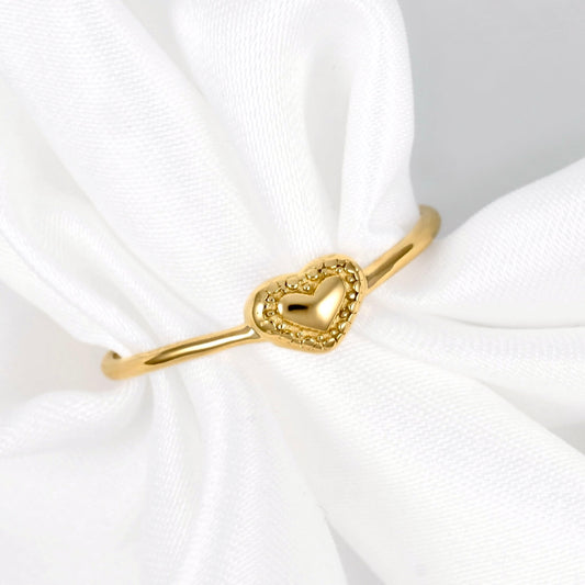 A gold ring with a milgrain heart-shaped  charm, displayed on a white fabric ring holder.