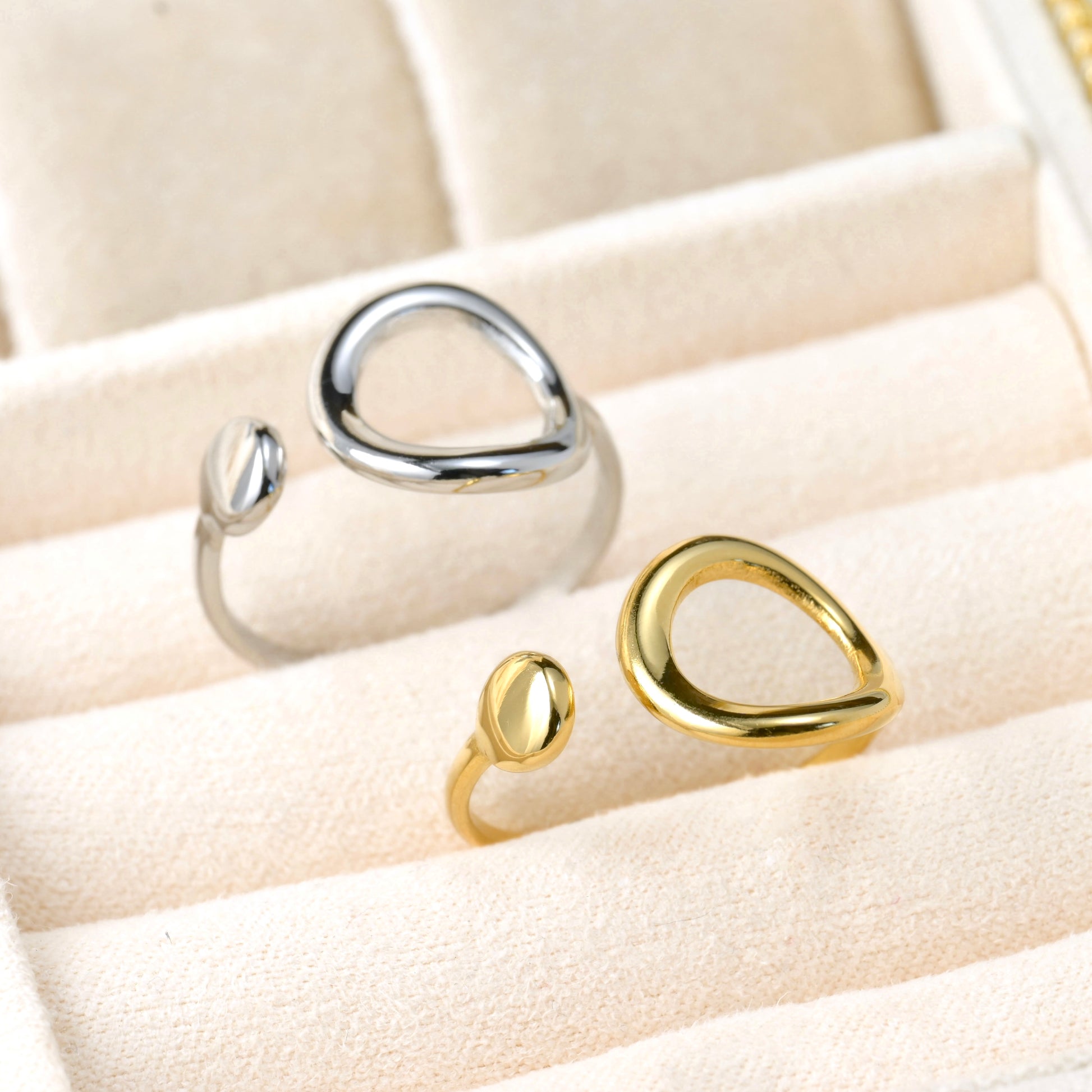 Two rings in silver and gold with asymmetrical circle designs on a beige cushioned surface