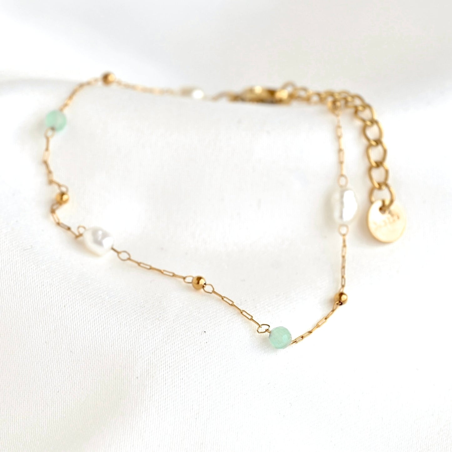 close up view of a delicate gold chain bracelet with small green and white beads, displayed on a soft white fabric background.