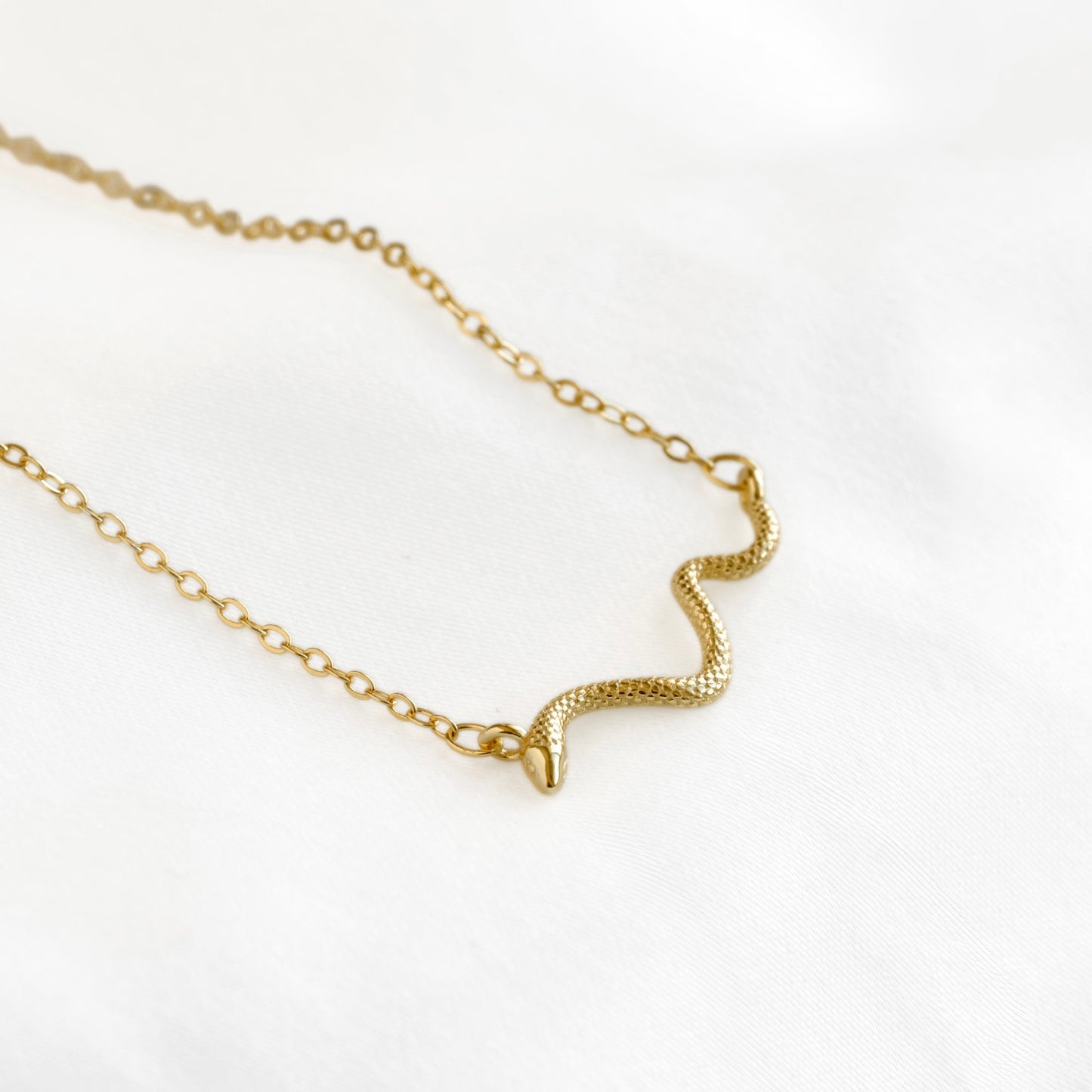 A delicate gold chain necklace featuring a small, textured snake pendant, displayed on a soft white fabric background.
