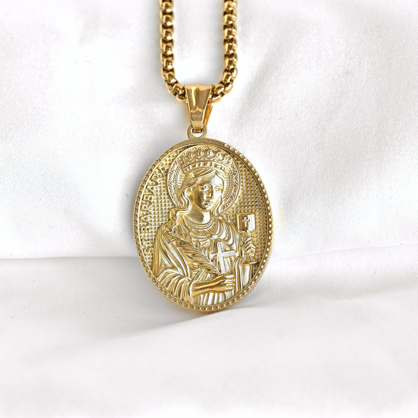 A gold oval religious pendant necklace featuring an embossed depiction of the Virgin Mary, suspended from a gold chain against a white fabric background.