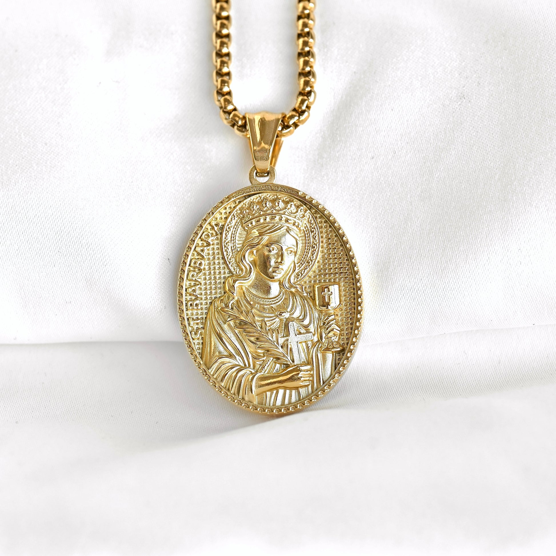A gold oval religious pendant necklace featuring an embossed depiction of the Virgin Mary, suspended from a gold chain against a white fabric background.