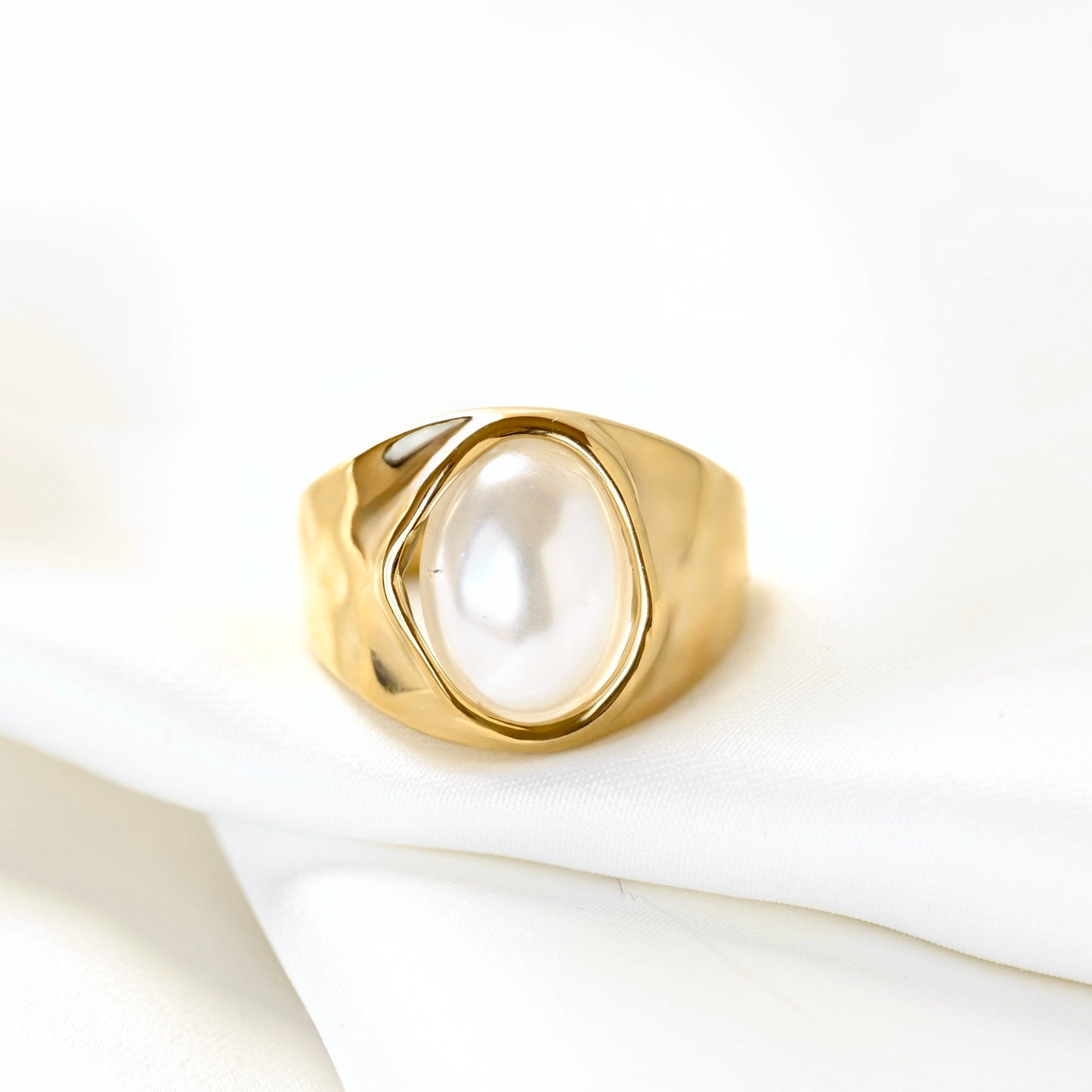 A gold signet-style ring featuring a large, smooth oval white pearl, resting on a soft white fabric surface.