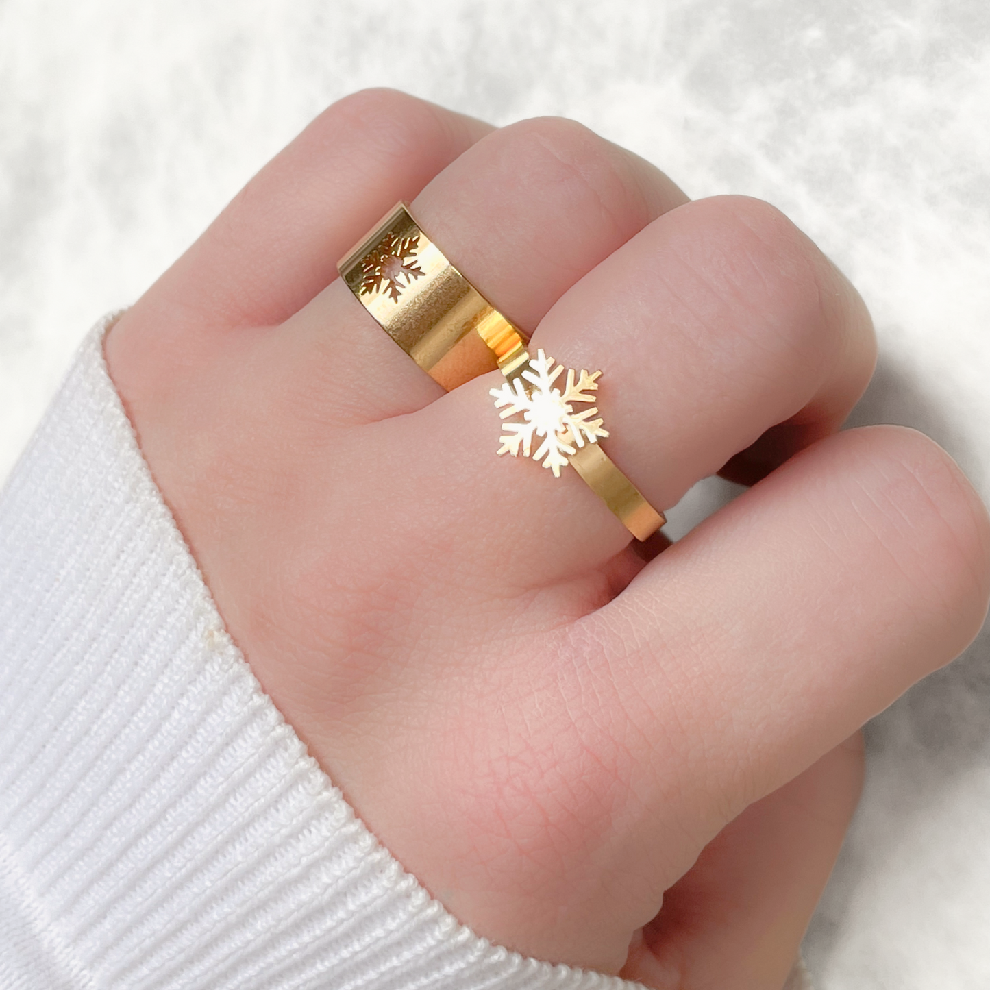 Gold rings with snowflake design, one flat slim with a large snowflake charm and one flat bold ring with snowflake cutout on a finger against a white background