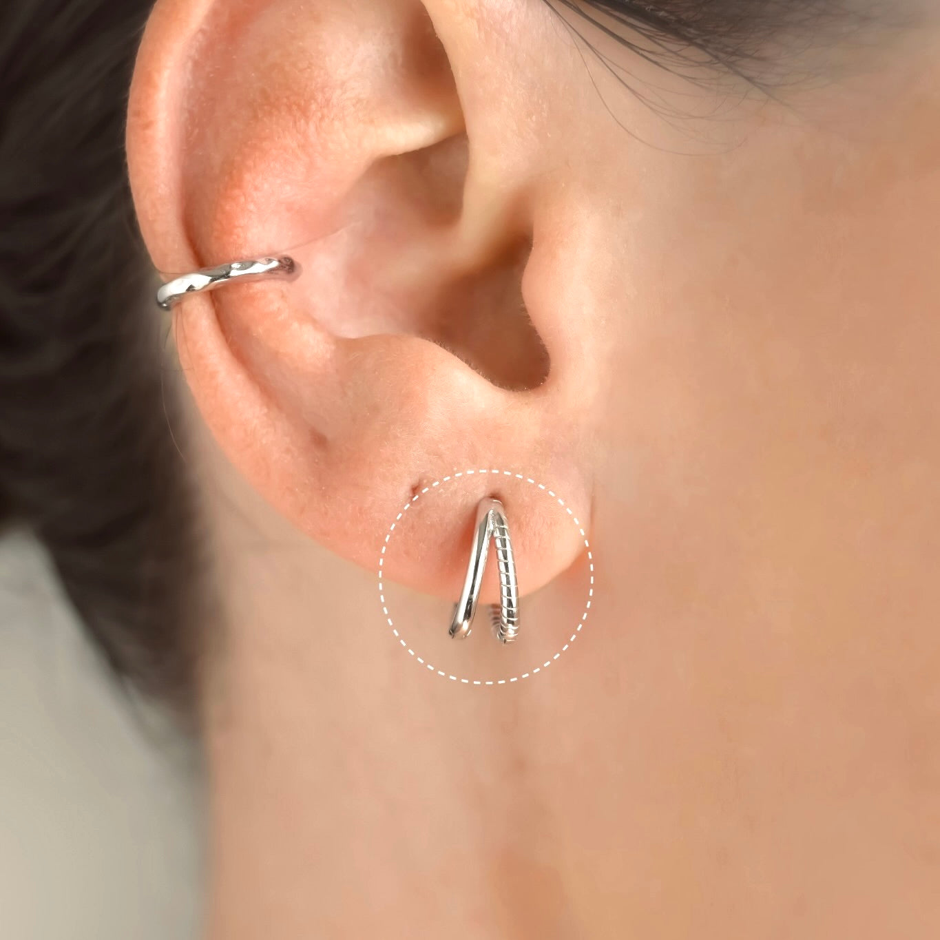 Close-up of an ear wearing a  layered silver hoop earring with plain and twisted bands with a blurred background