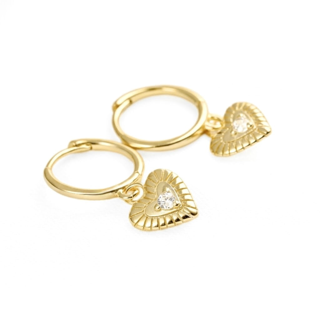 A pair of gold hoop earrings with textured heart charms, each featuring a small clear gemstone in the center, set against a plain white background.