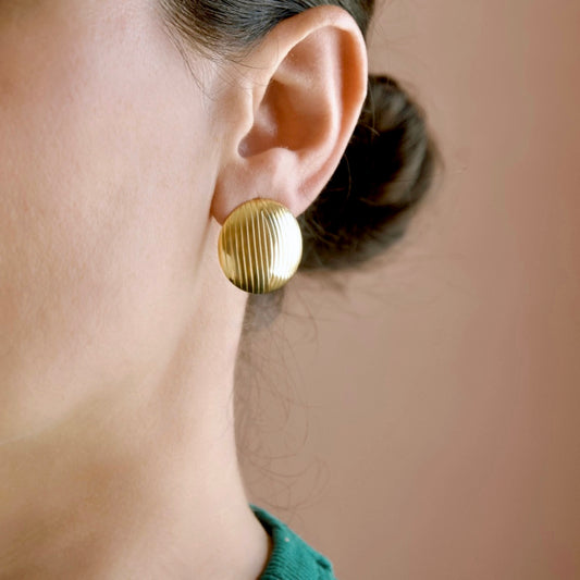 A close-up side profile of a person wearing a single round gold earring with vertical ridges, set against a soft warm beige background.