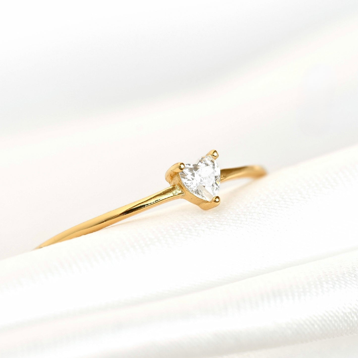 A yellow gold solitaire ring featuring a heart-shaped clear gemstone, resting on a white satin fabric surface.