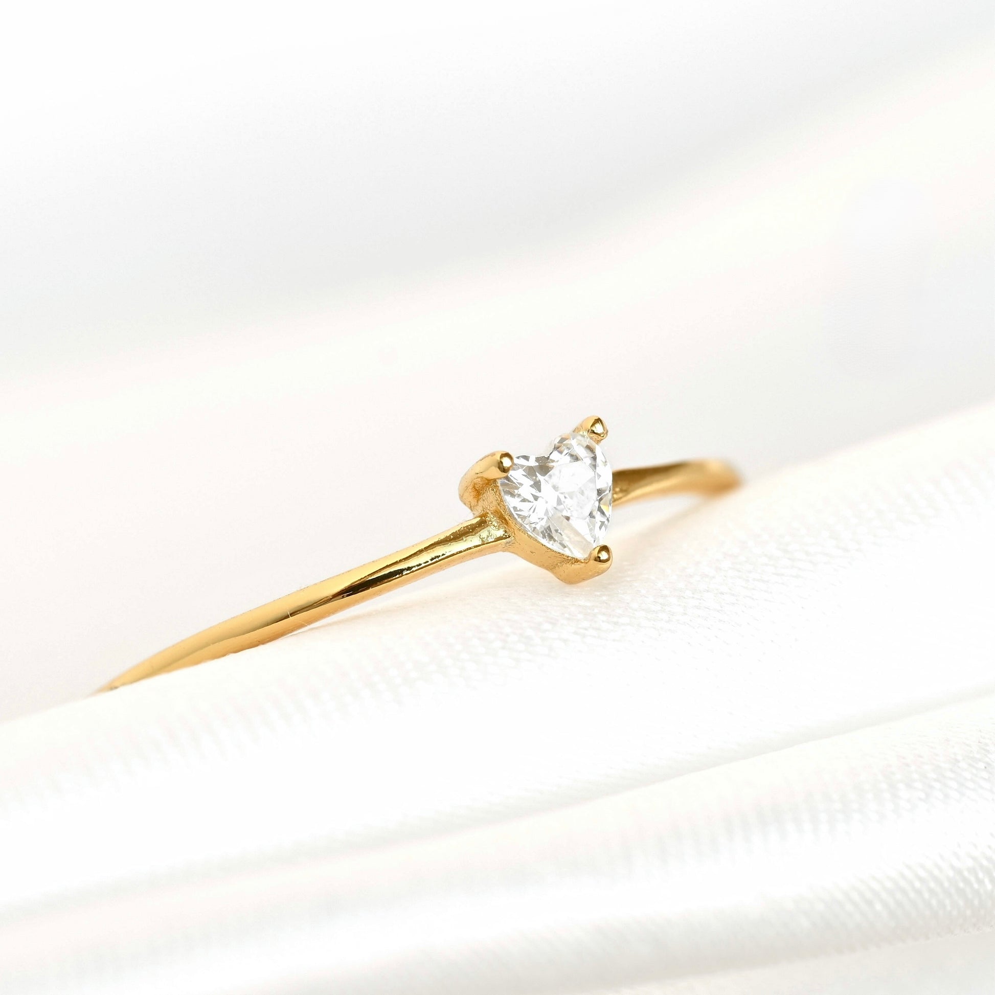 A yellow gold solitaire ring featuring a heart-shaped clear gemstone, resting on a white satin fabric surface.