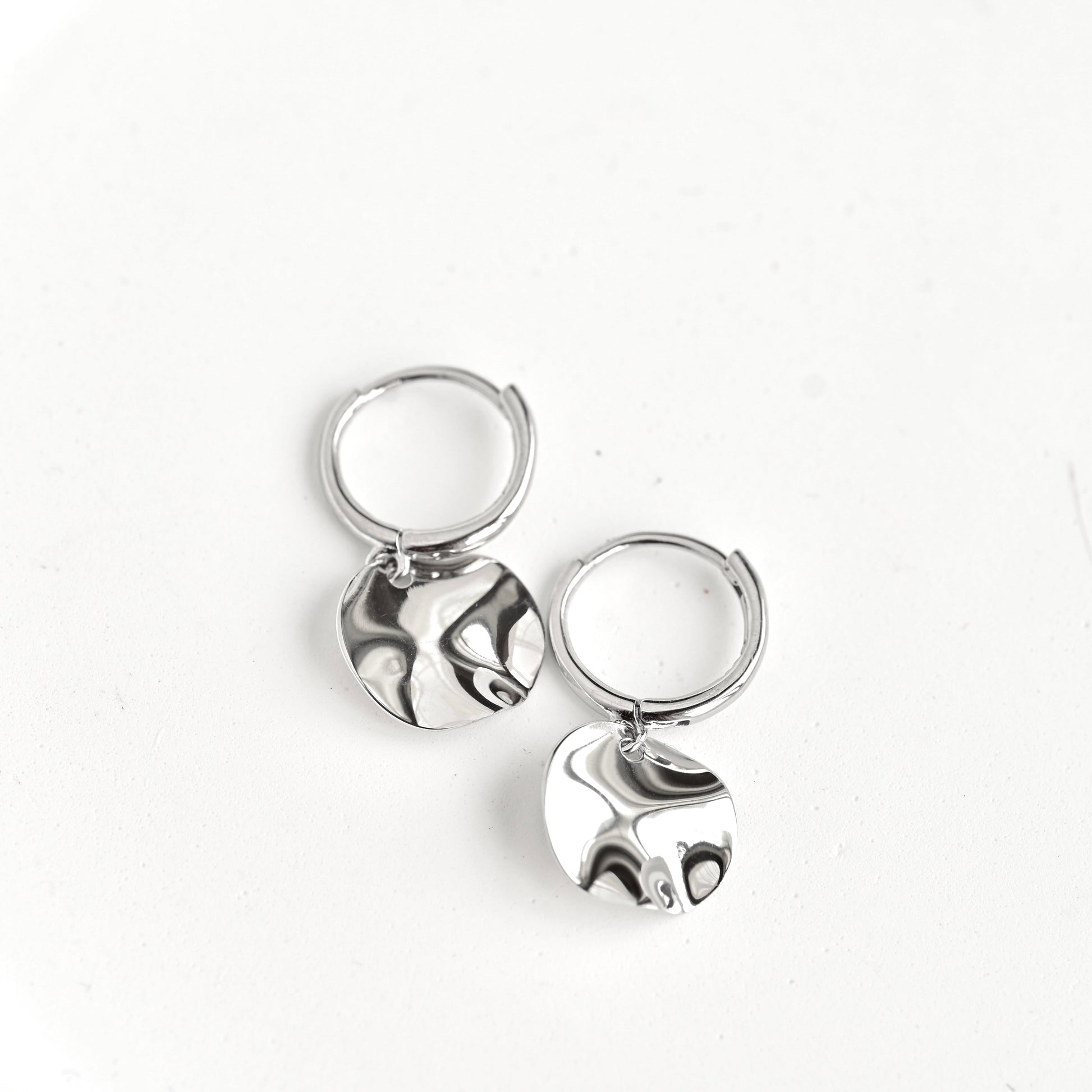 A pair of silver huggie hoop earrings with flat, textured circular dangles, displayed on a clean white background.