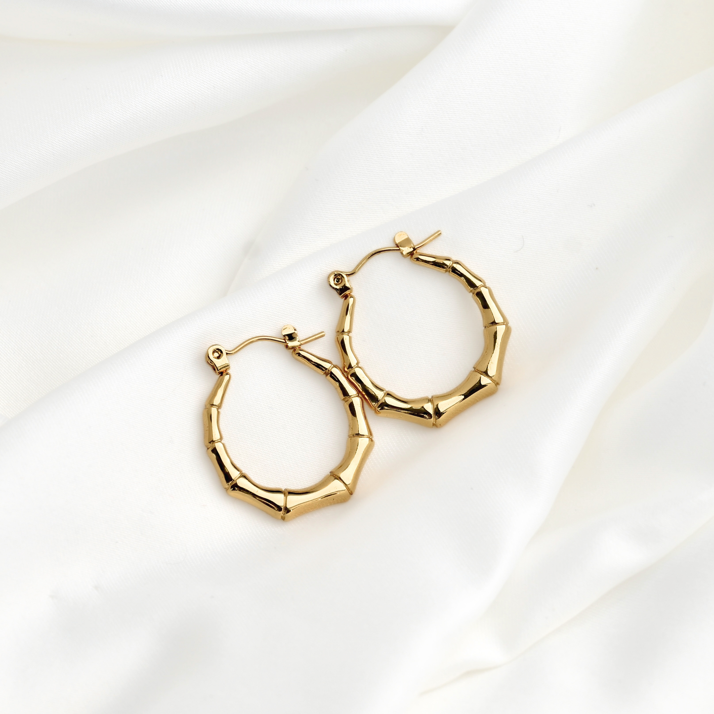 A pair of gold hoop earrings with a textured, segmented design resembling bamboo joints, displayed on a draped white satin fabric background.