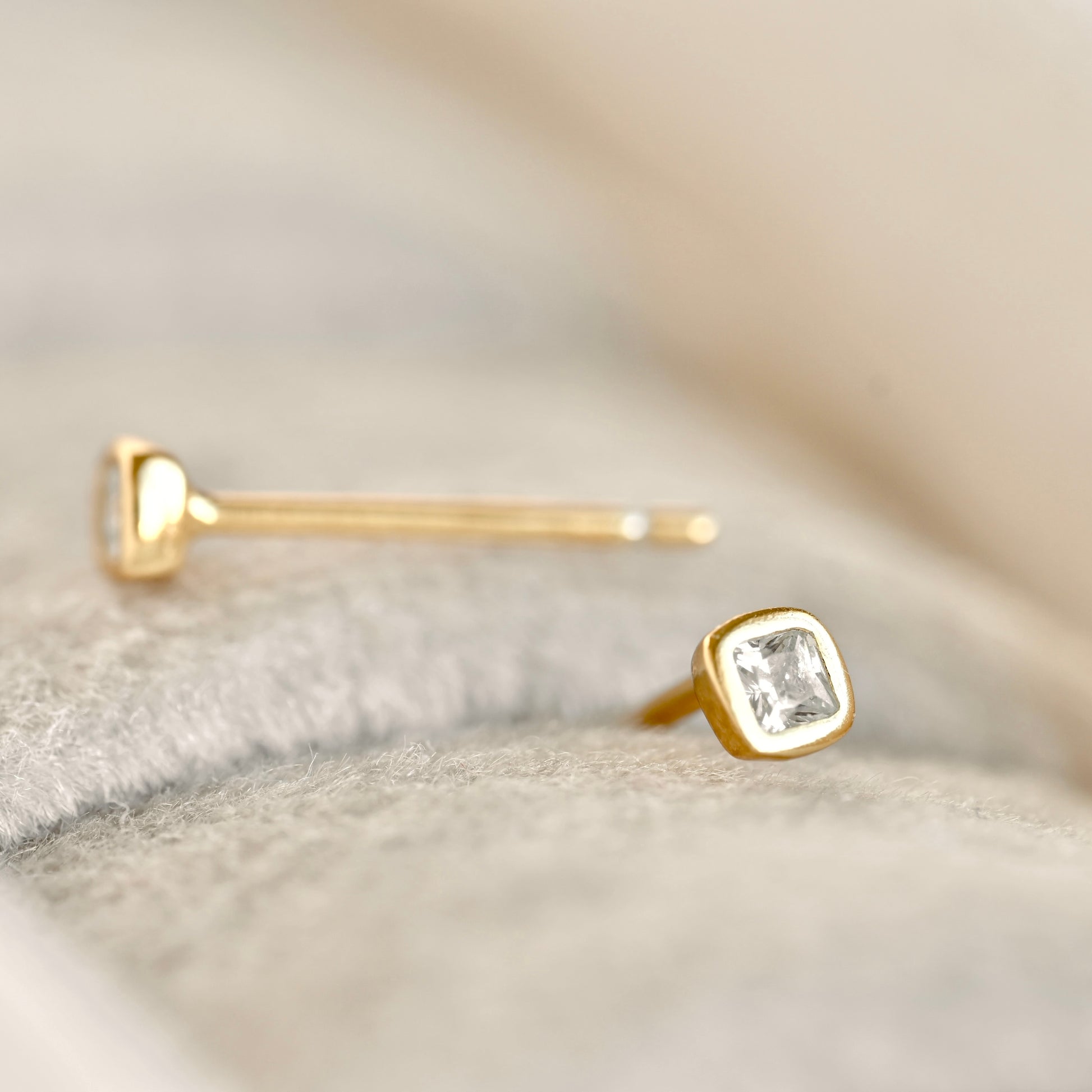 A pair of gold stud earrings with clear rectangular gemstones, resting on a soft, textured light gray fabric surface.