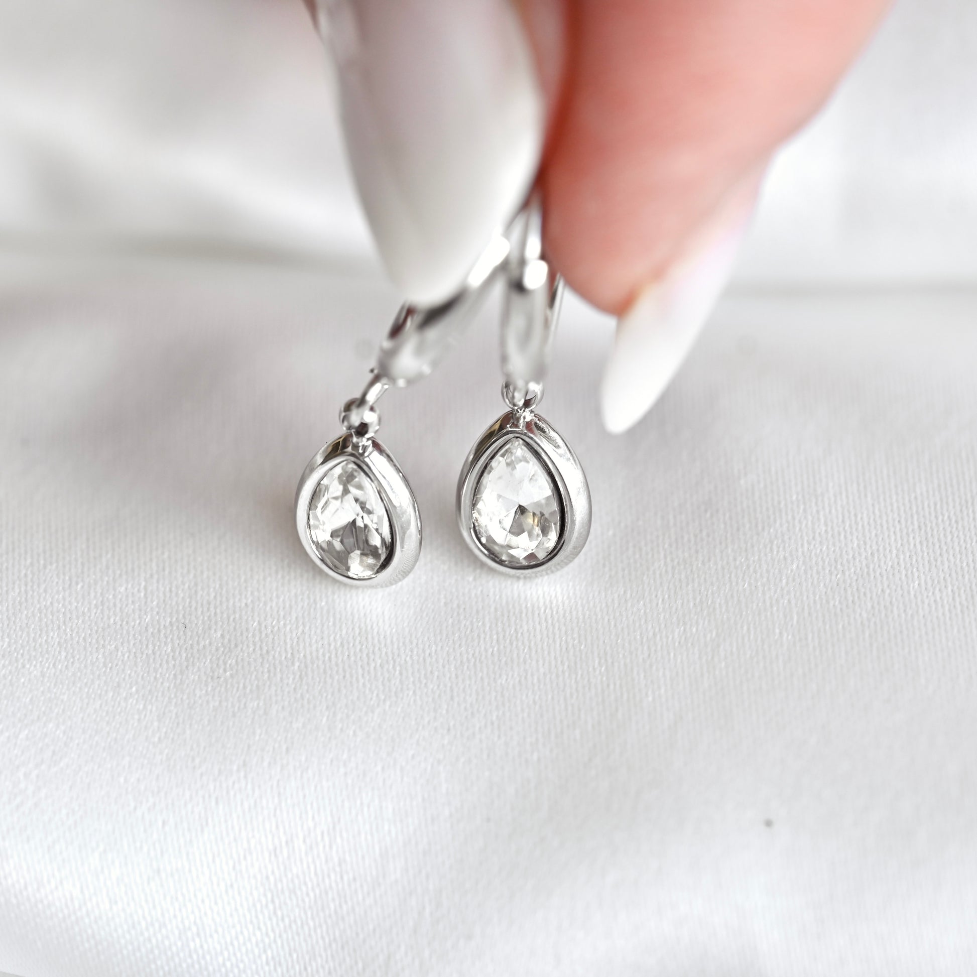 A close-up of a hand with white manicured nails holding a pair of silver drop earrings with teardrop-shaped clear crystals against a white fabric background.