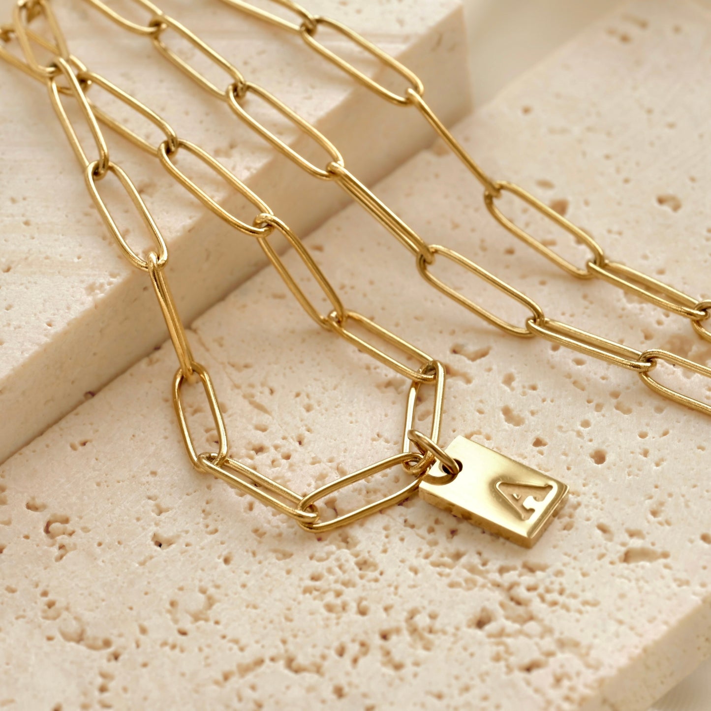 Gold paperclip necklace with a initial tag pendant on a textured beige surface
