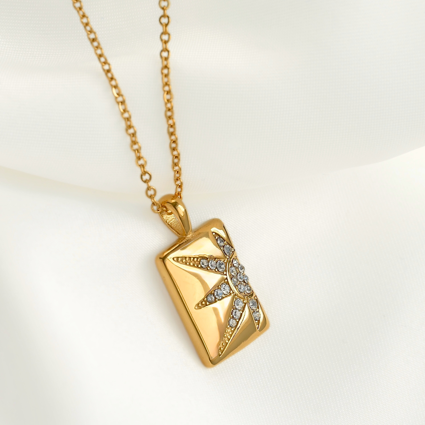 A gold rectangular pendant necklace featuring a half sun design paved with small clear stones, displayed on a soft white fabric background.