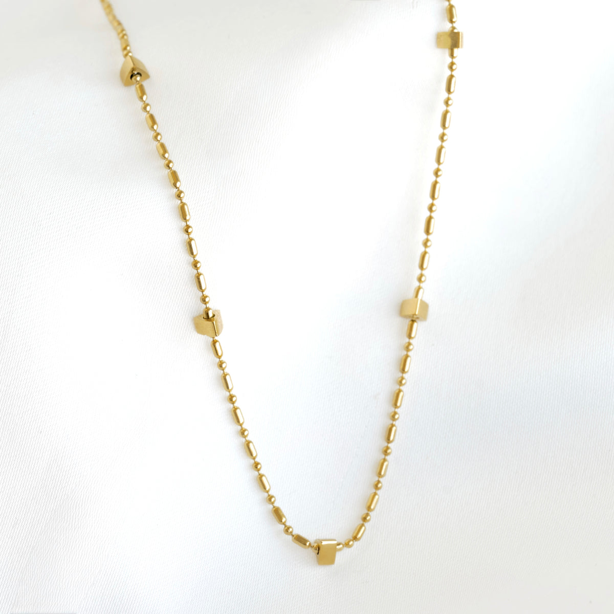 A delicate gold chain necklace featuring small, flat geometric charms in the shape of hearts, squares, and triangles spaced along the chain, displayed on a soft white fabric background.
