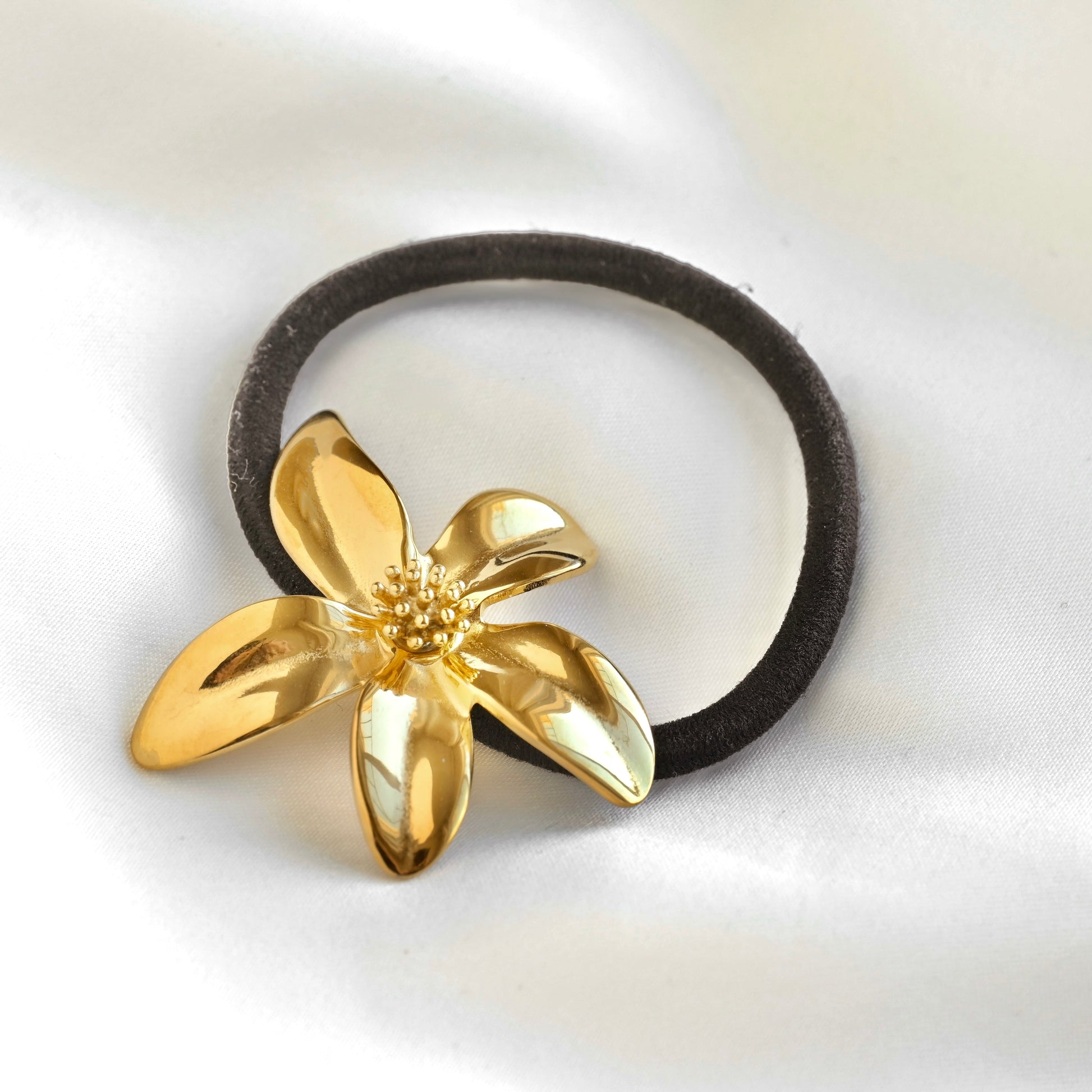 A black elastic hair tie featuring a shiny gold flower charm, displayed on a soft white satin fabric background.