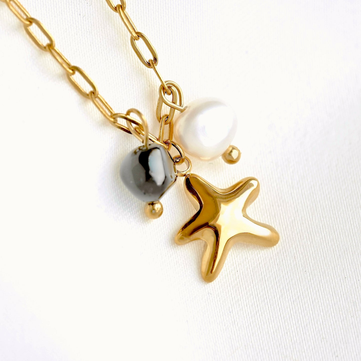 Close up view of gold paperclip chain bracelet with a starfish and pearl charm, featuring a polished gold starfish and a white pearl bead, alongside a small dark stone, on a white textured background.