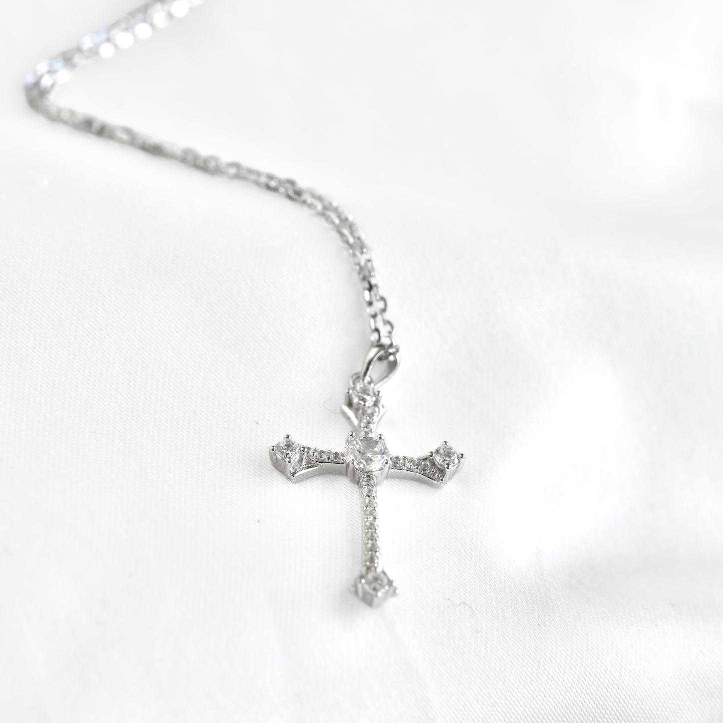 A silver cross pendant necklace adorned with clear crystals, resting on a white fabric background.
