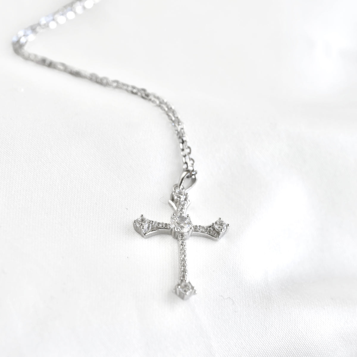 A silver cross pendant necklace adorned with clear crystals, resting on a white fabric background.