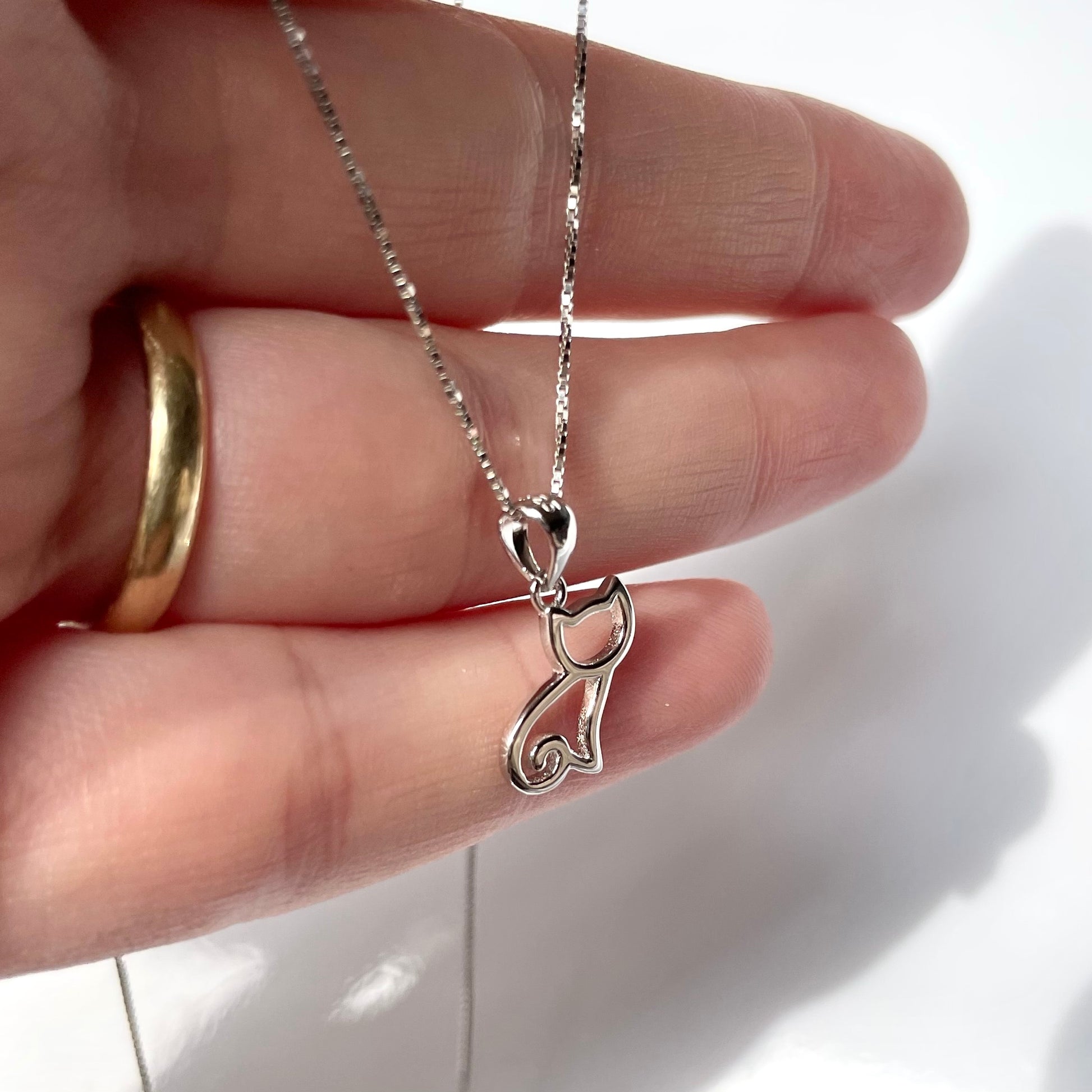 Silver cat charm necklace held by a hand with a gold ring on a white background
