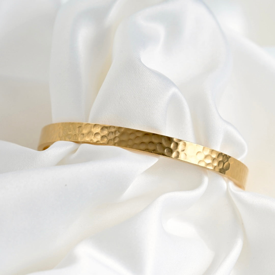 A hammered gold bangle bracelet displayed on draped white satin fabric.