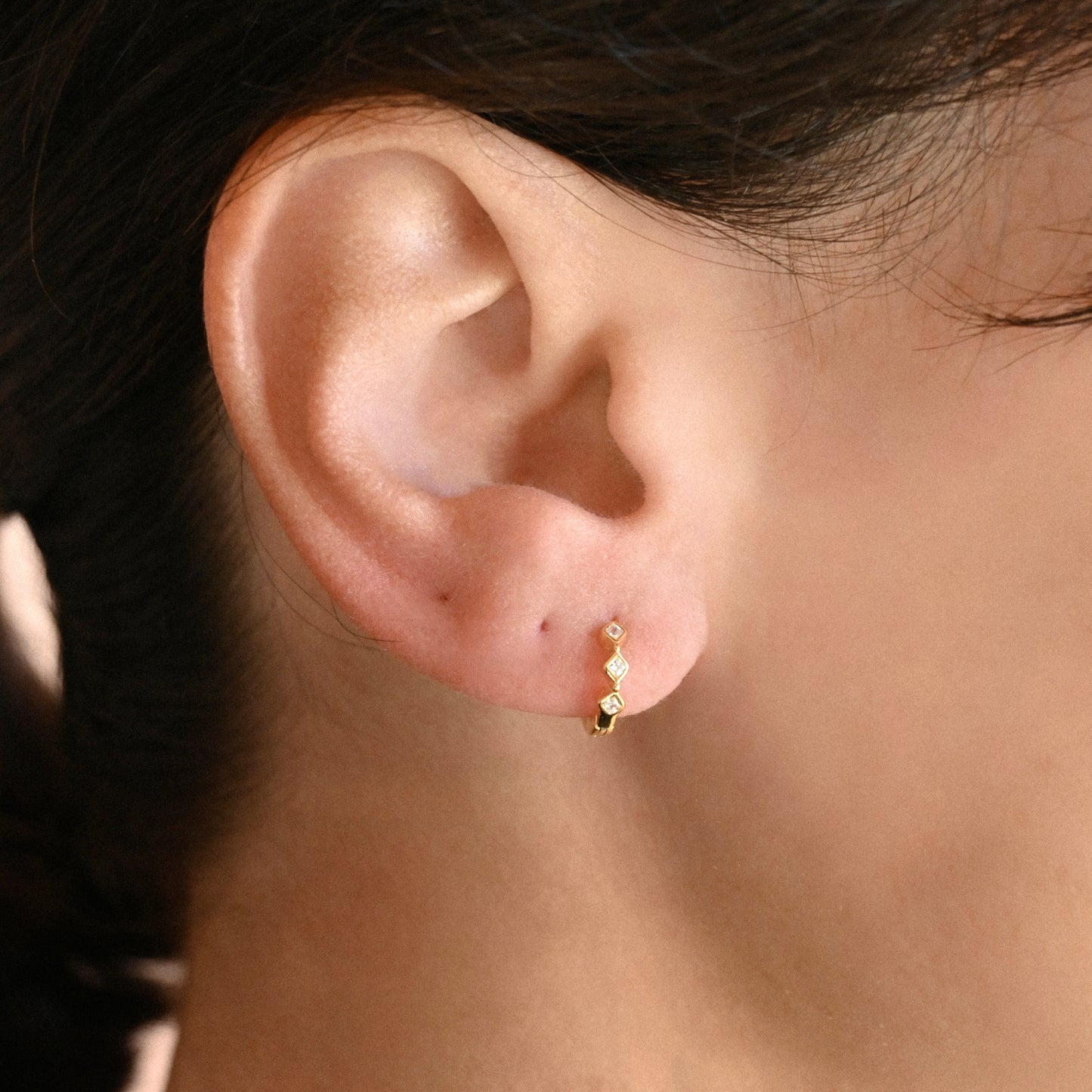 A close-up of a person's ear wearing a small gold huggie hoop earring with three tiny rhombus-shaped clear stones in a bezel setting