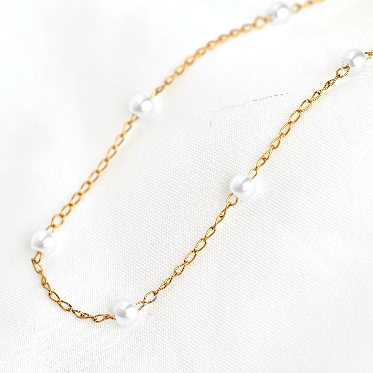 A delicate gold chain necklace with evenly spaced, small white pearl beads, displayed on a textured white fabric background.