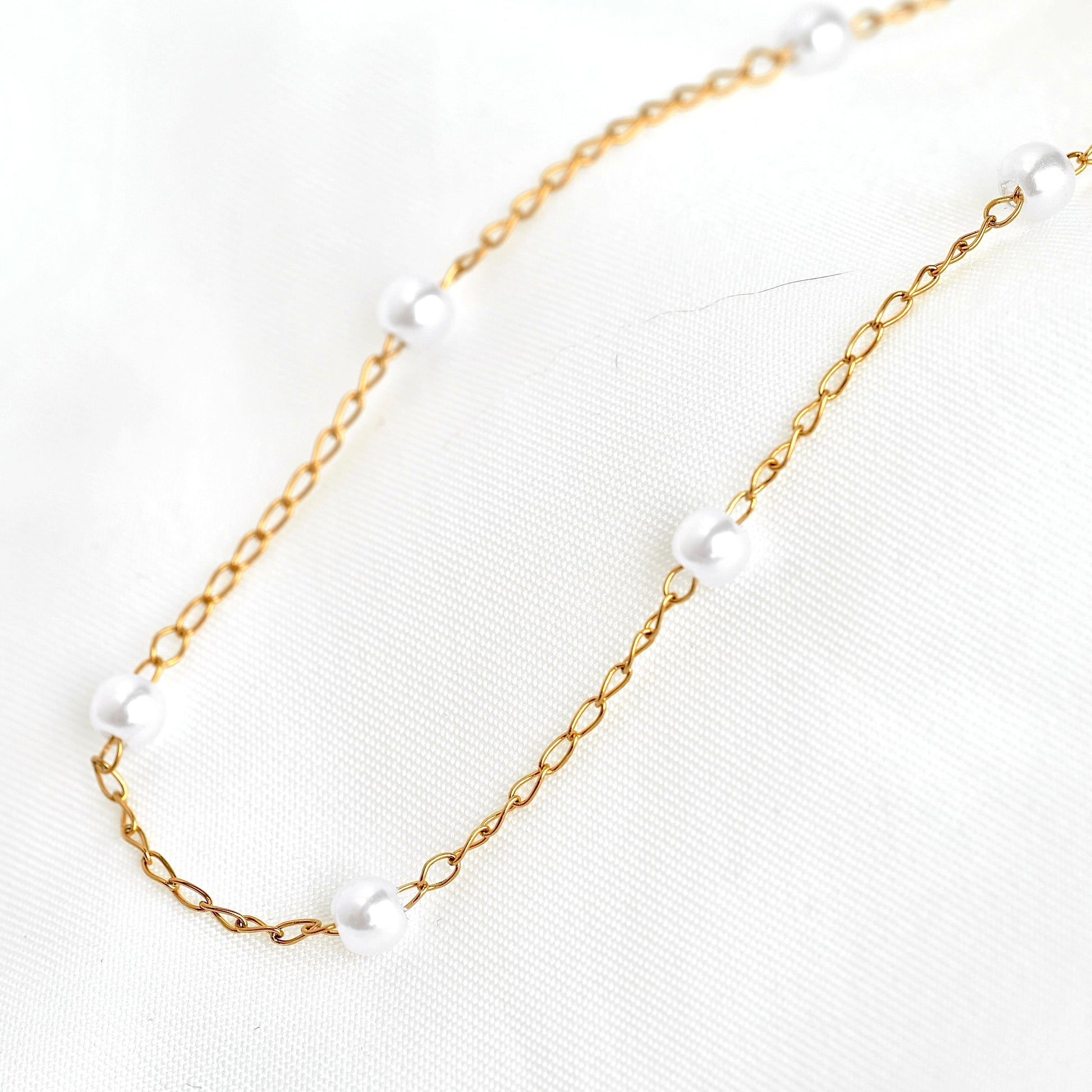 A delicate gold chain necklace with evenly spaced, small white pearl beads, displayed on a textured white fabric background.