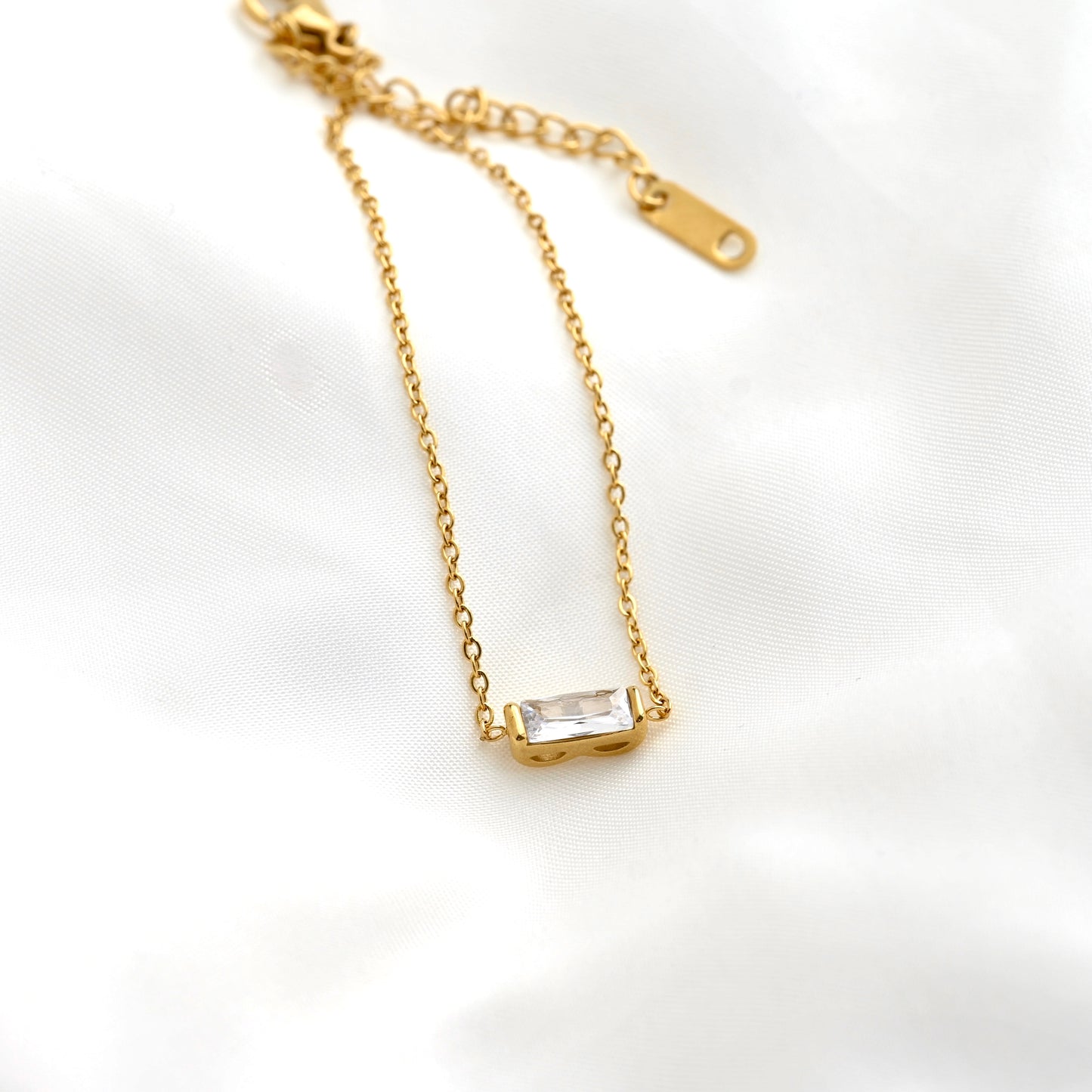 A delicate gold chain bracelet with a small baguette stone connector, displayed on a soft white fabric background.