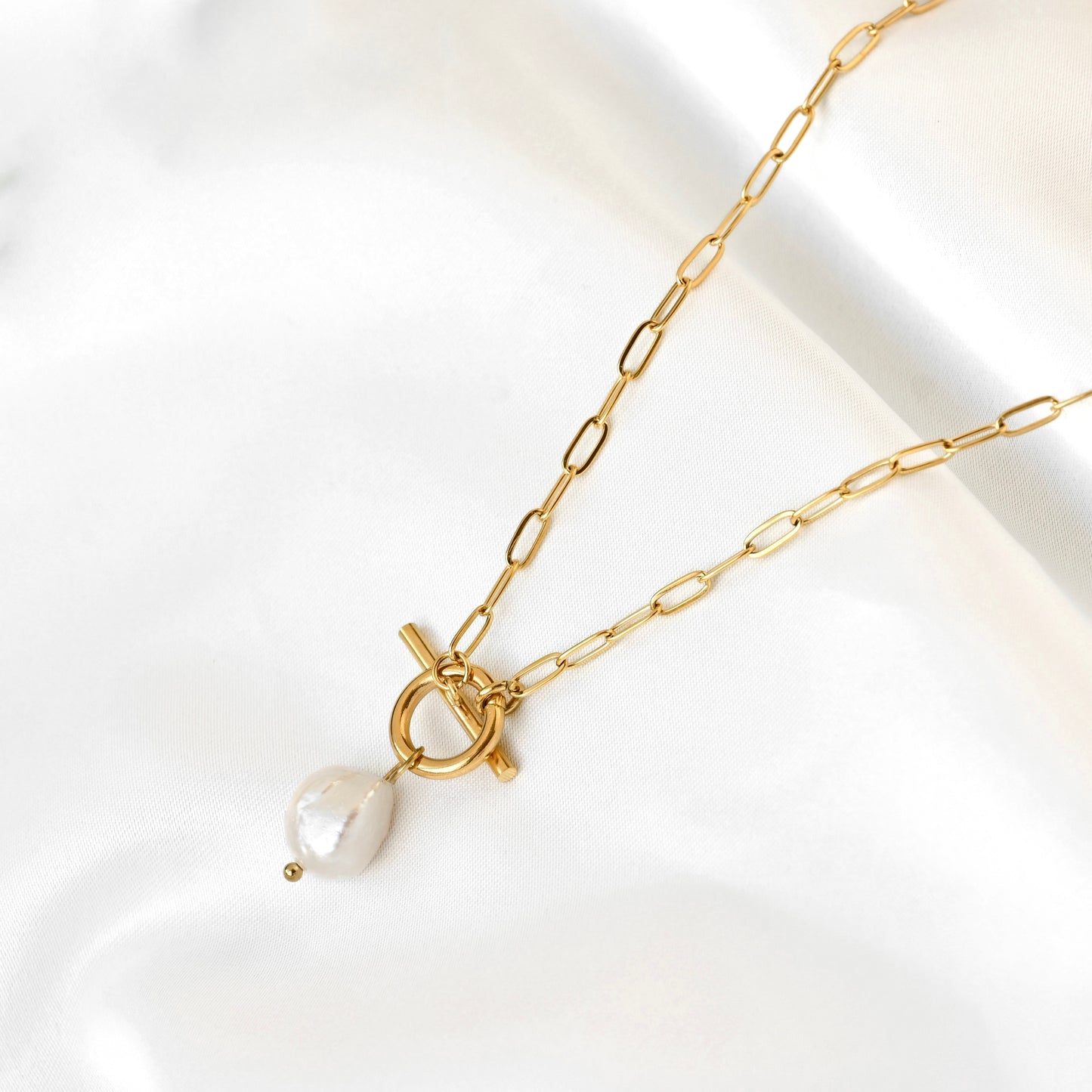 A gold paperclip chain necklace with a toggle clasp and a small white baroque pearl pendant, displayed on a soft white satin fabric.