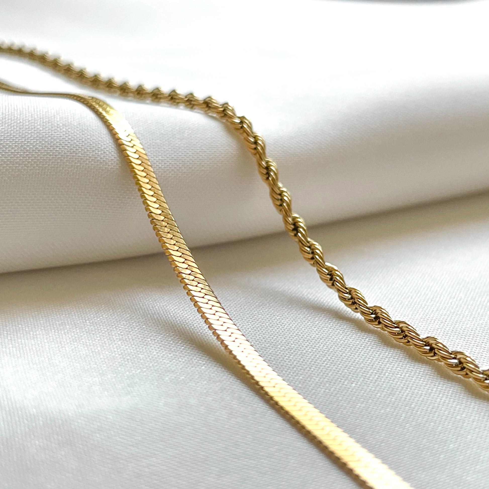 Angled front view of layered gold chain necklace with the flat herringbone reflecting light beautifully on a white fabric background