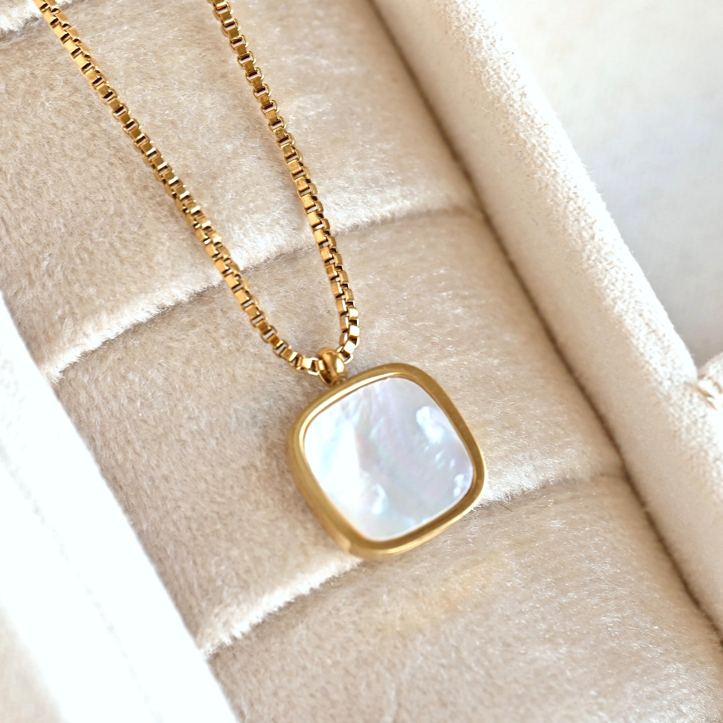 A gold chain necklace with a square iridescent mother-of-pearl pendant, displayed inside a cream-colored velvet jewelry box.