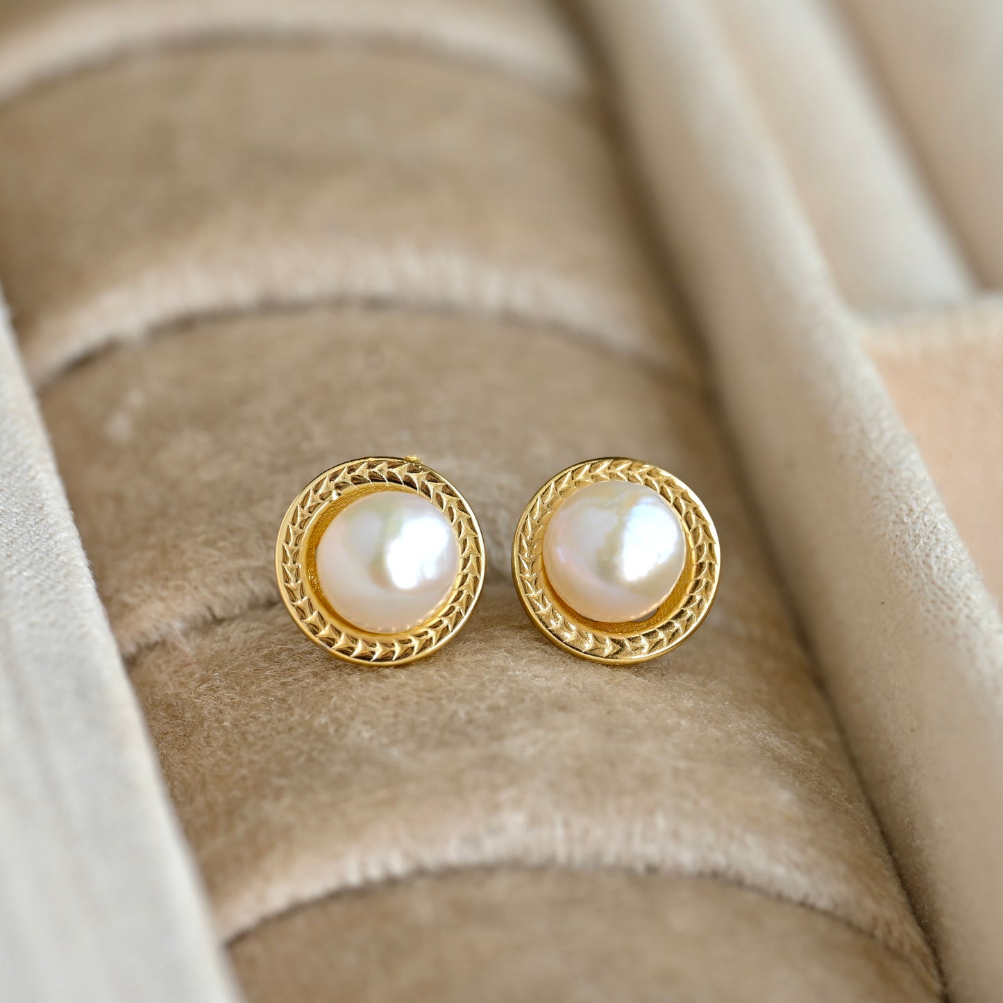 A pair of vintage style gold pearl stud earrings with round white pearls and a braided gold border, displayed on a beige velvet jewelry ring holder.