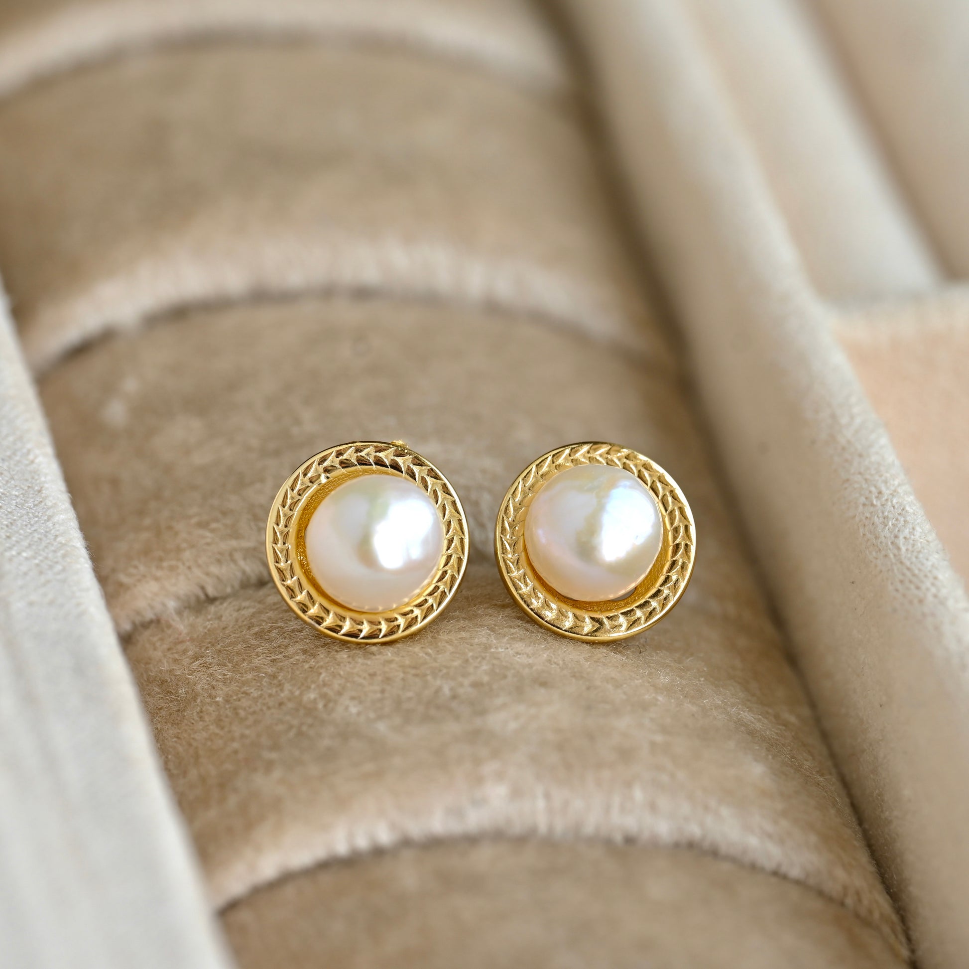 A pair of vintage style gold pearl stud earrings with round white pearls and a braided gold border, displayed on a beige velvet jewelry ring holder.