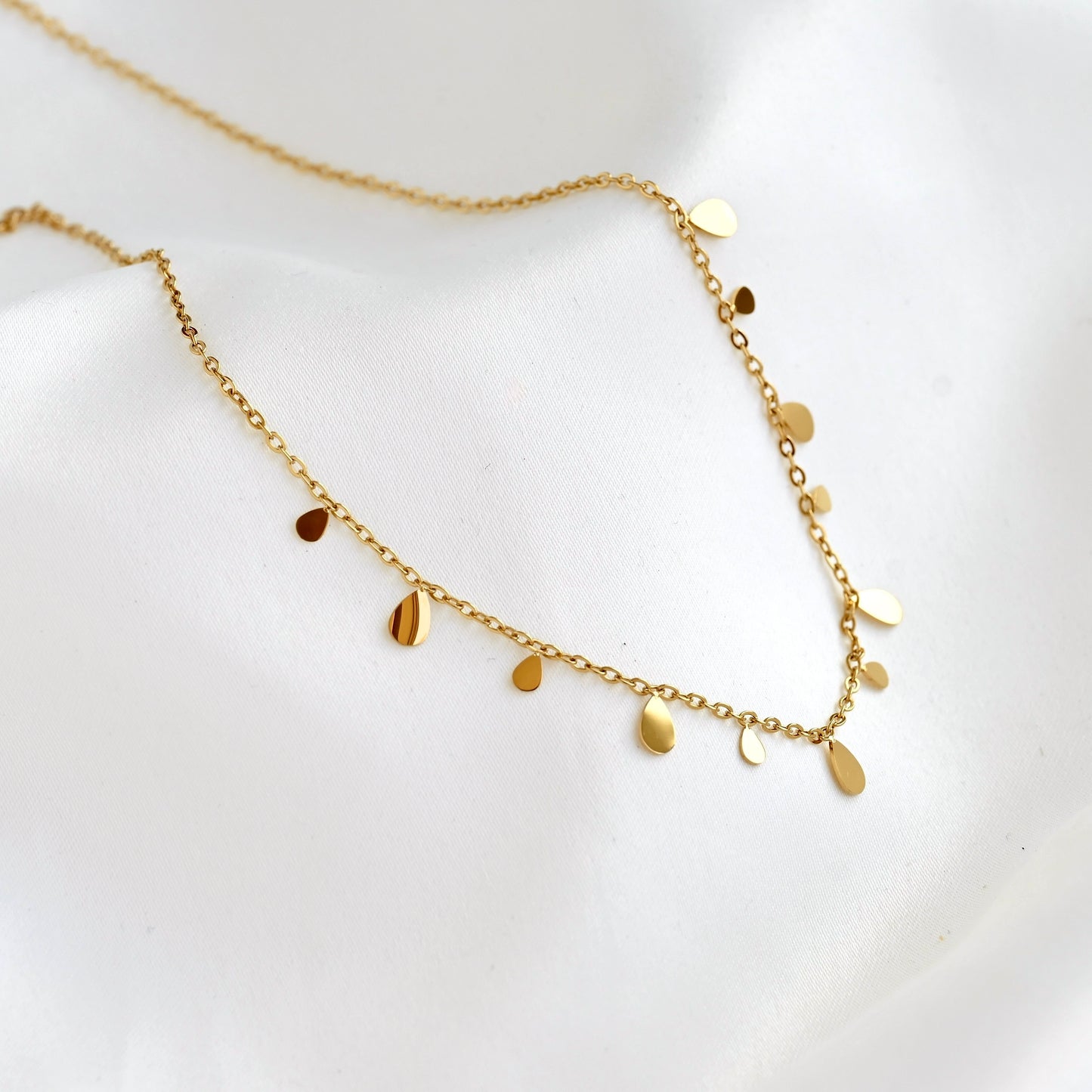 A delicate gold chain necklace with small flat teardrop-shaped discs, displayed on a soft white fabric background.