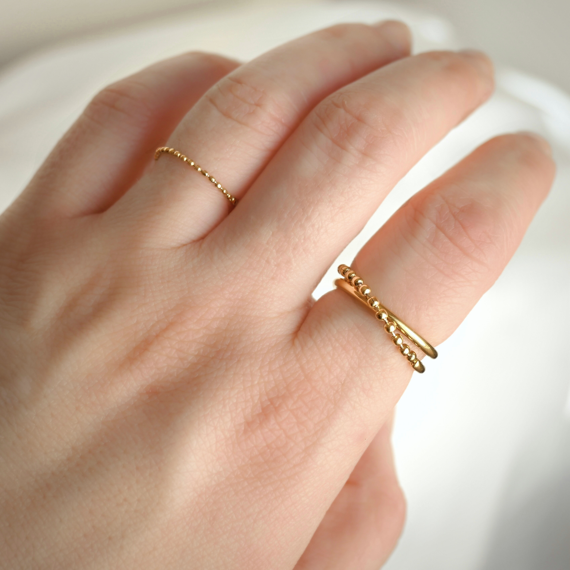 A close-up of a hand wearing two delicate gold beaded rings, with one ultra thin chain ring on the ring finger and one textured beaded band on the index finger, set against a soft white background.