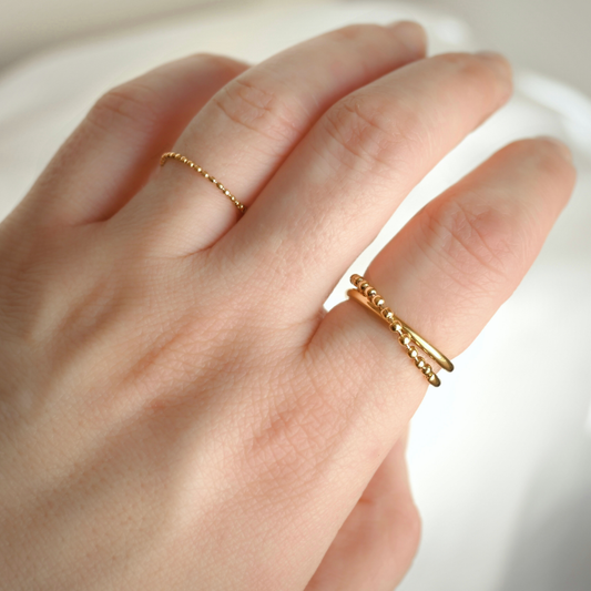 A close-up of a hand wearing two delicate gold beaded rings, with one ultra thin chain ring on the ring finger and one textured beaded band on the index finger, set against a soft white background.