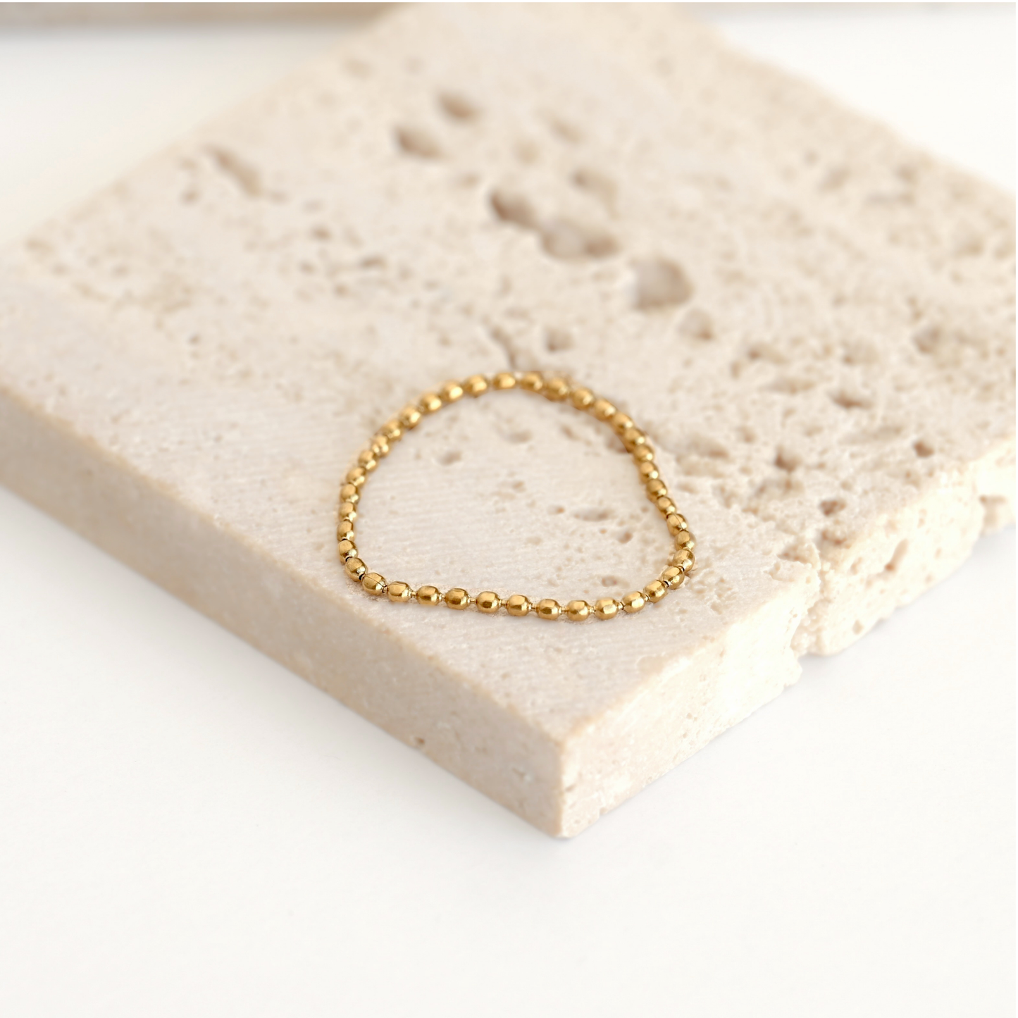 A delicate gold beaded bracelet resting on a textured beige stone tile against a clean white background.
