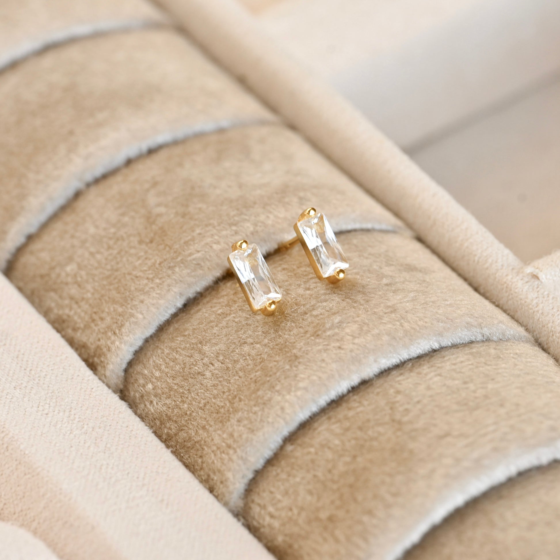 Gold baguette stud earrings with clear gemstones on a beige textured surface