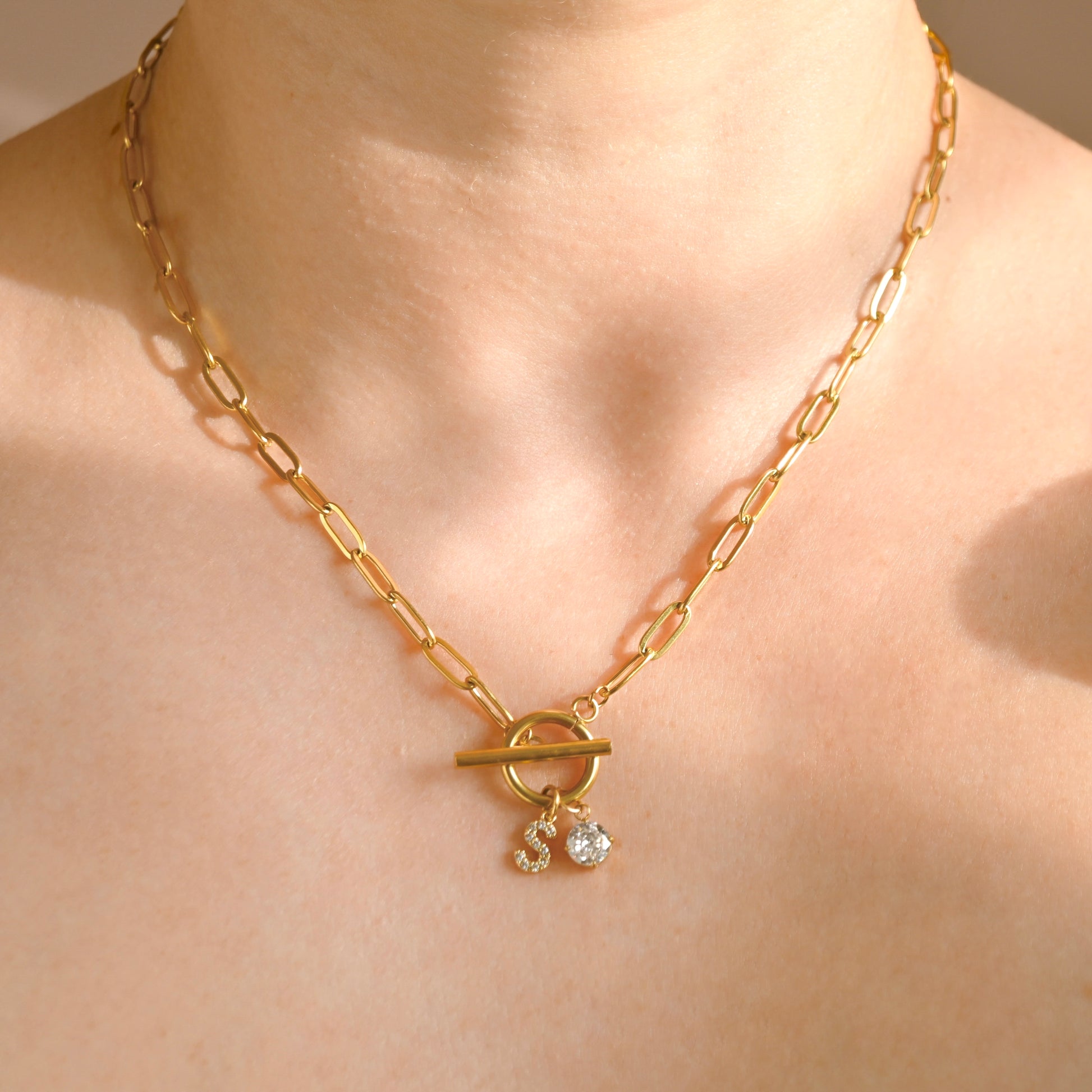 A gold paperclip chain necklace with a toggle clasp and two small clear crystal charms, shown worn on a model's neck.