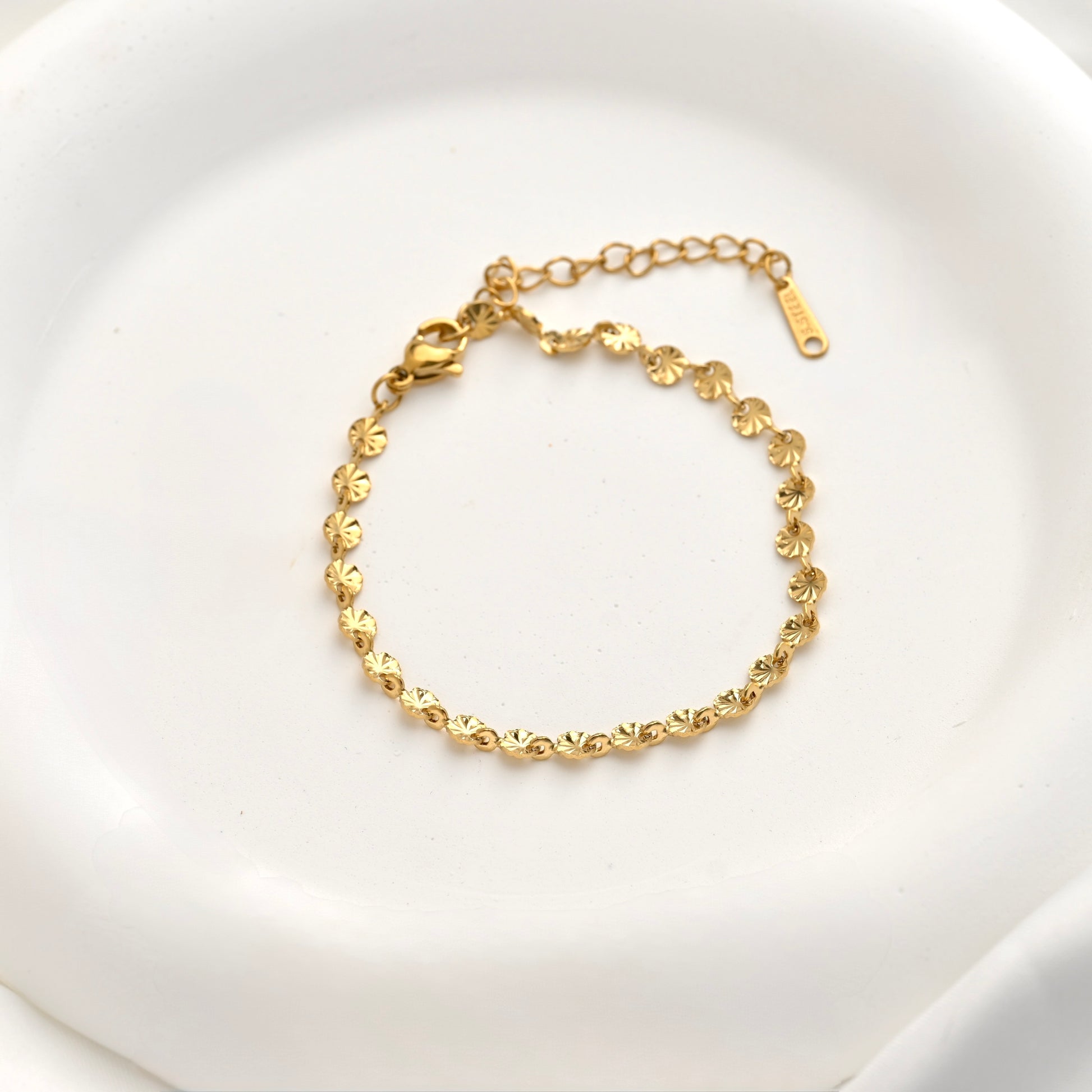 A delicate gold chain bracelet with textured disc links, displayed on a white ceramic dish.